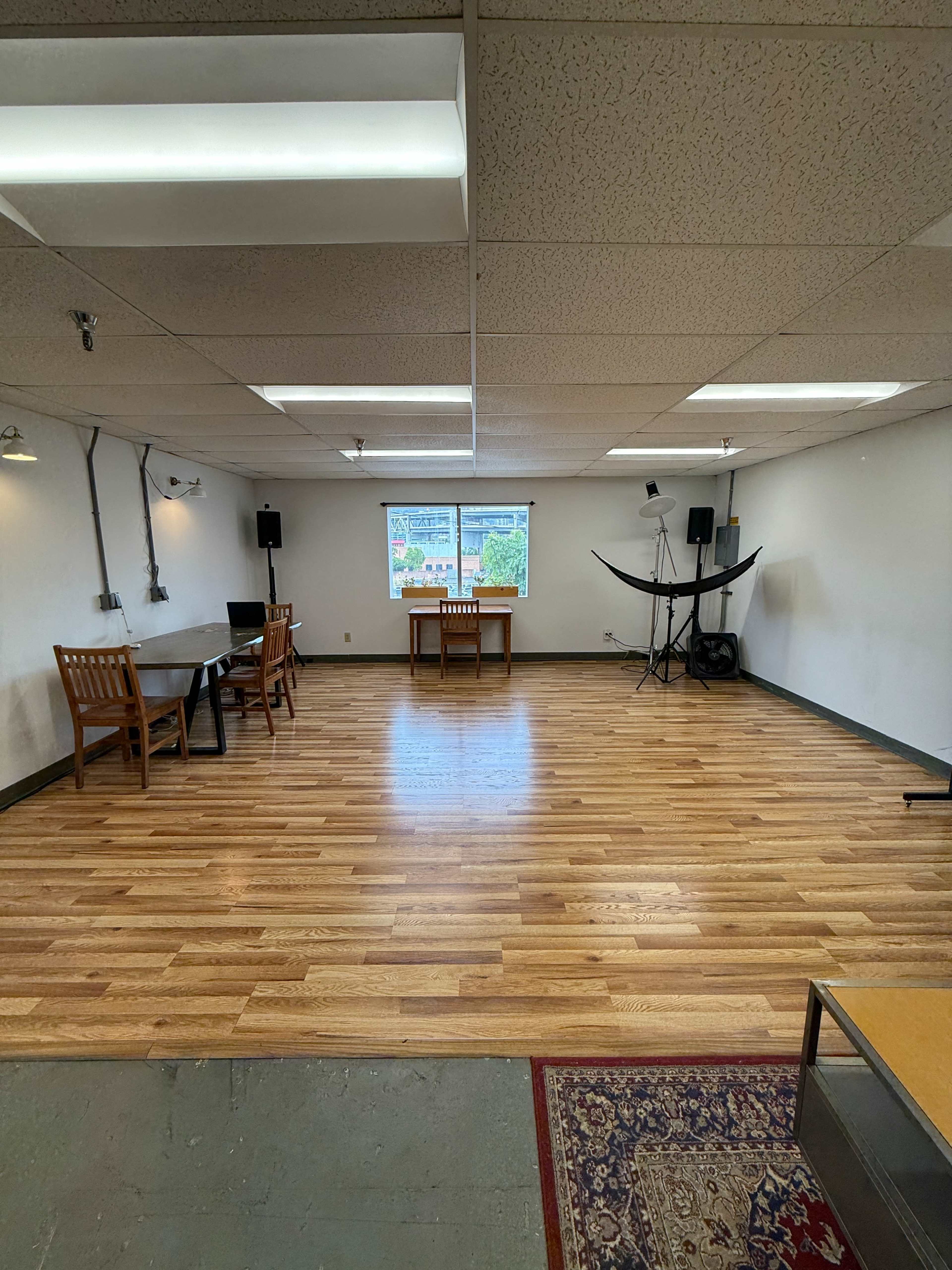 The image shows a spacious, empty room with wooden flooring, two tables with chairs, and equipment set up in the corners.