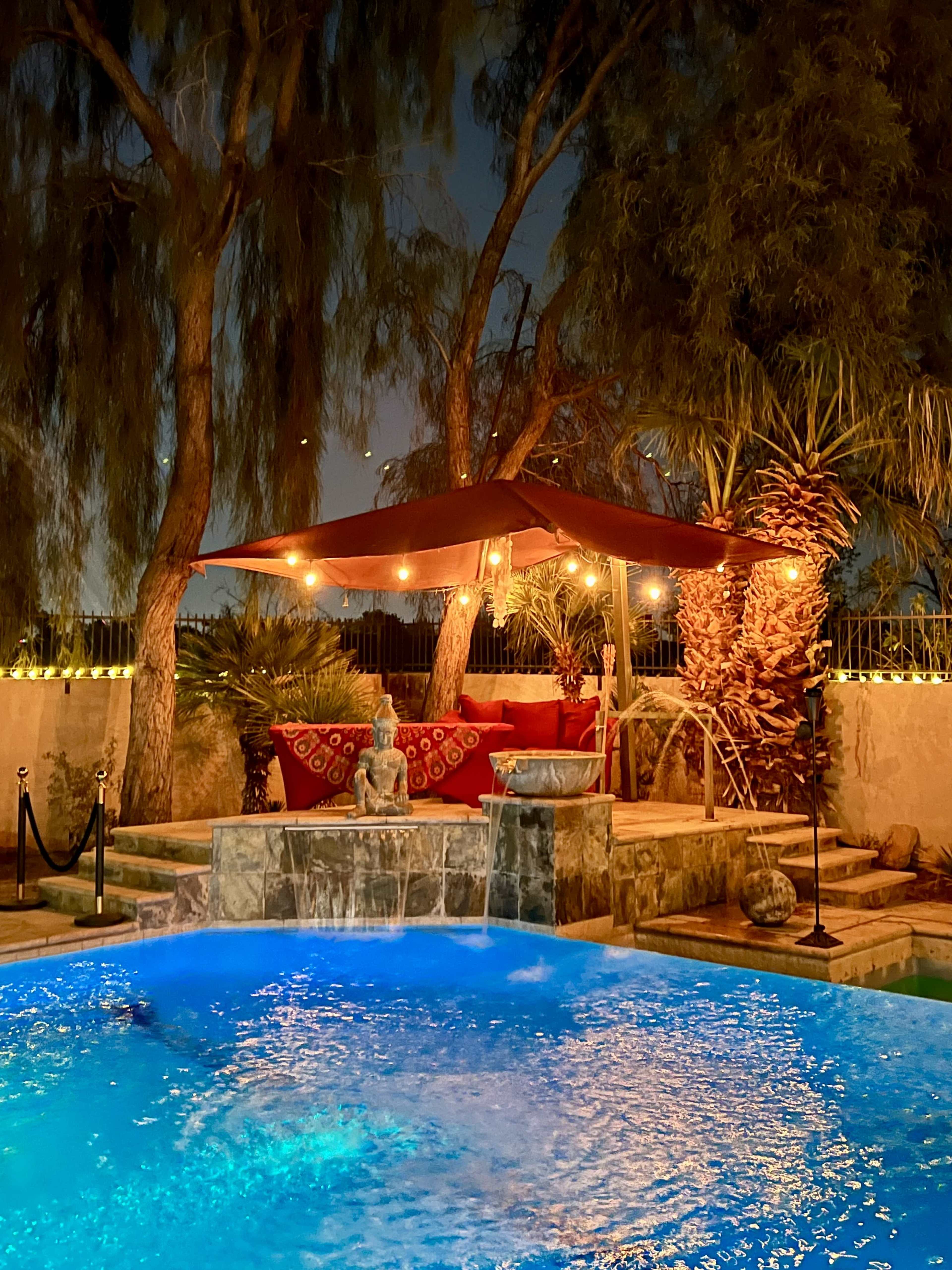 The image shows a well-lit outdoor area featuring a pool, a stone waterfall, a decorative statue, and a shaded seating area under a canopy.