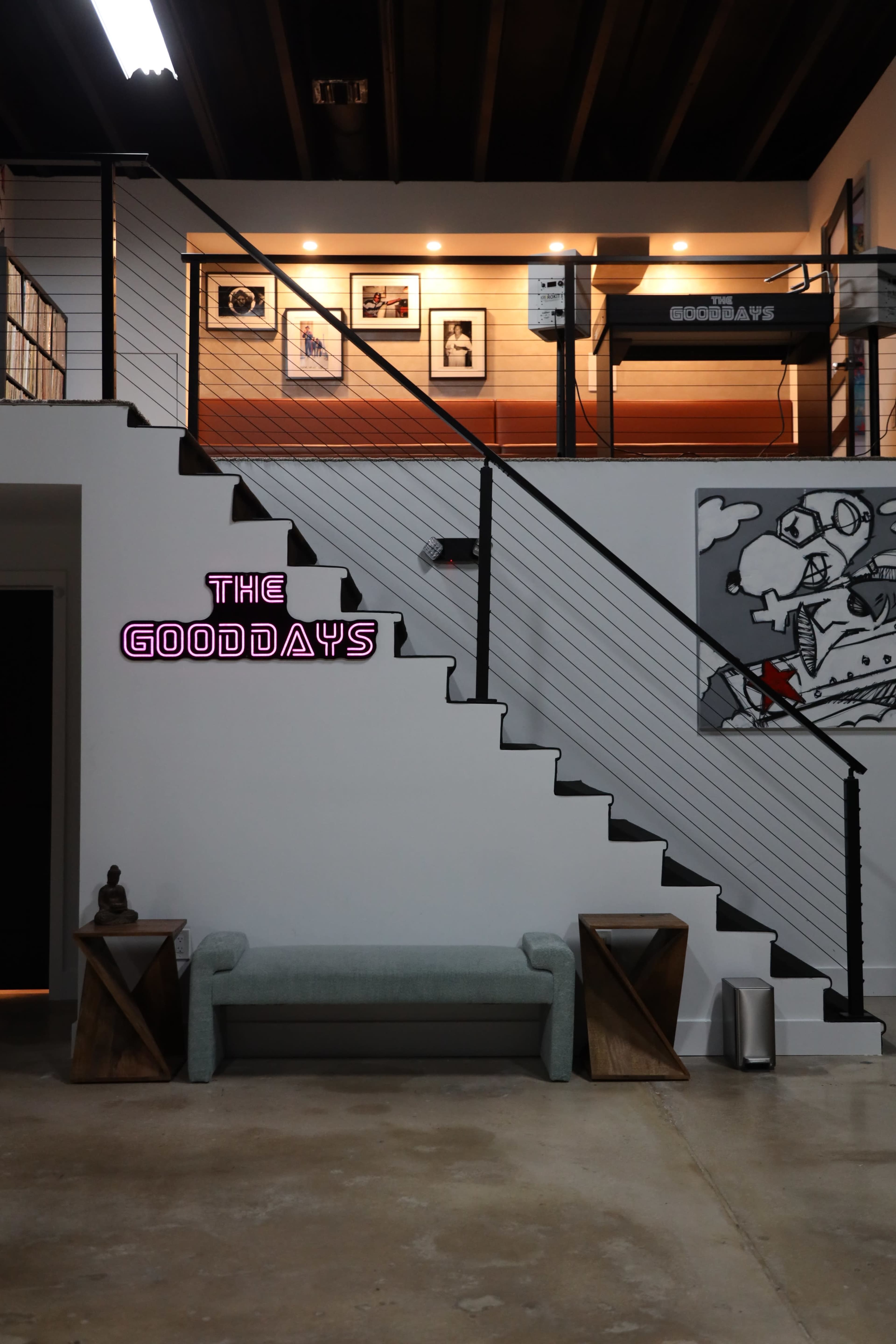 The image shows a modern interior with a staircase featuring a neon sign that reads "THE GOODDAYS," alongside a bench and decorative elements in a minimalist design.