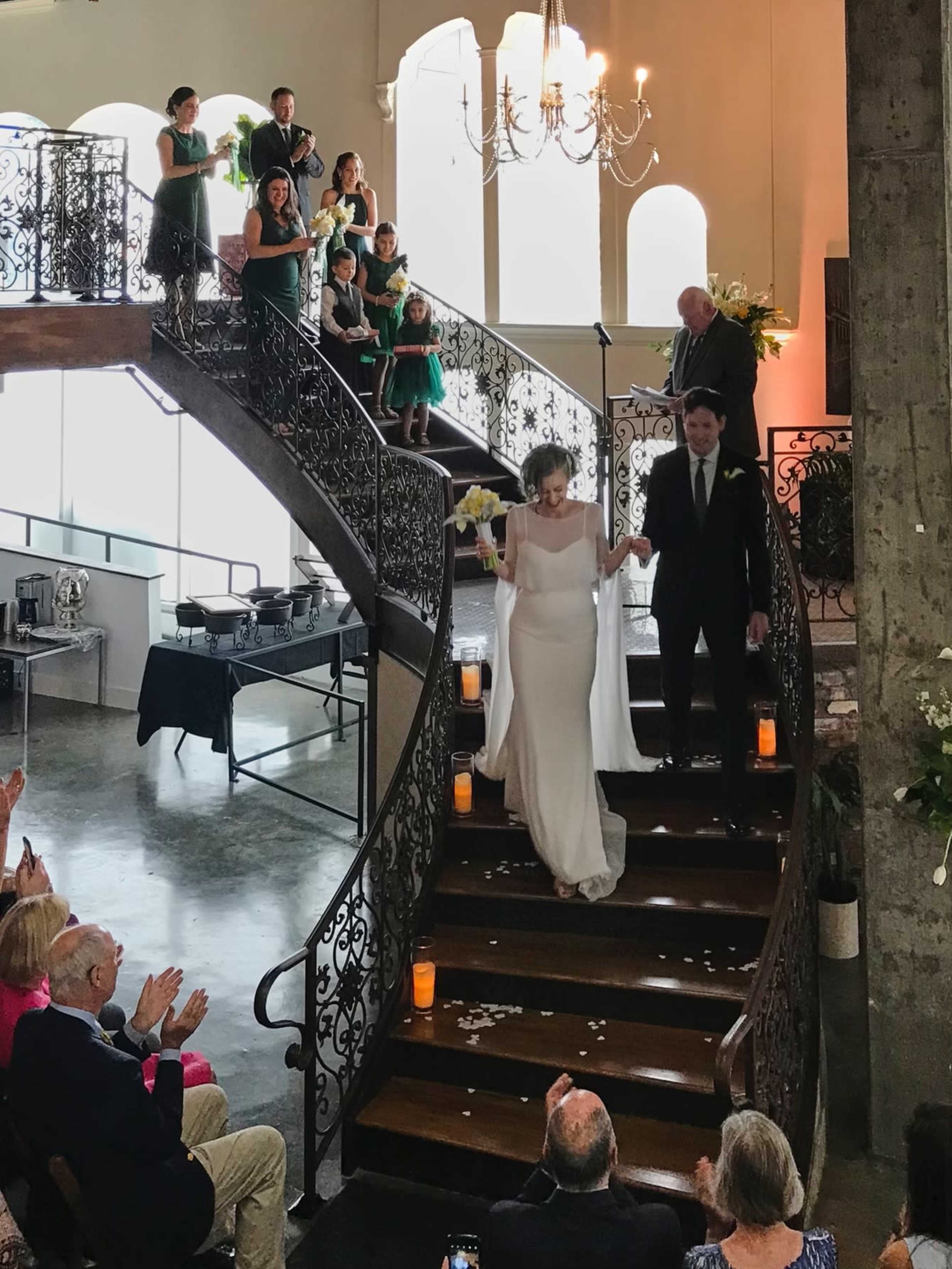 A bride and groom descend a staircase in a decorated venue while guests applaud from below.