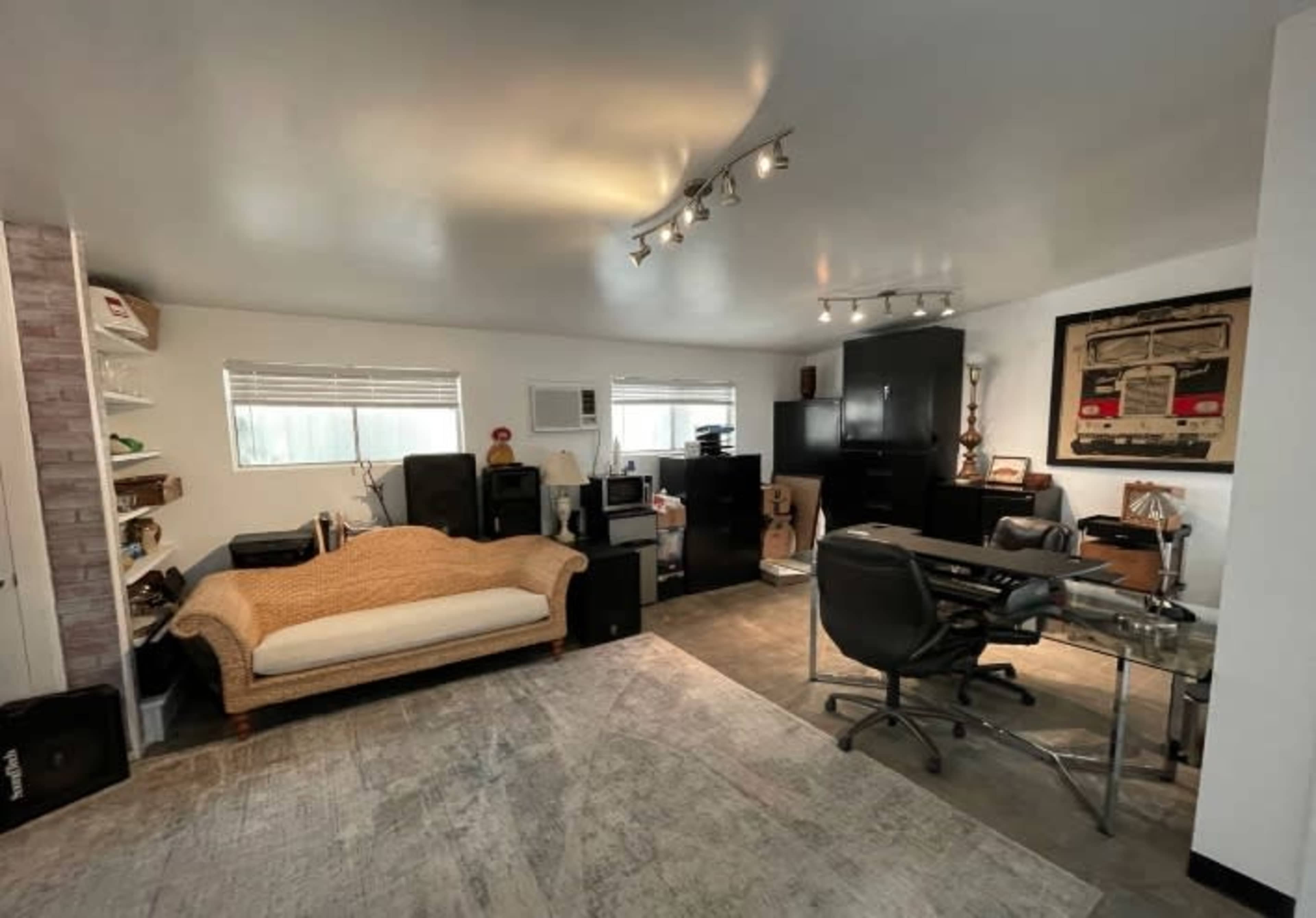 A modern home office space featuring a glass desk, a black office chair, a light-colored sofa, and various audio equipment and storage units along the walls.