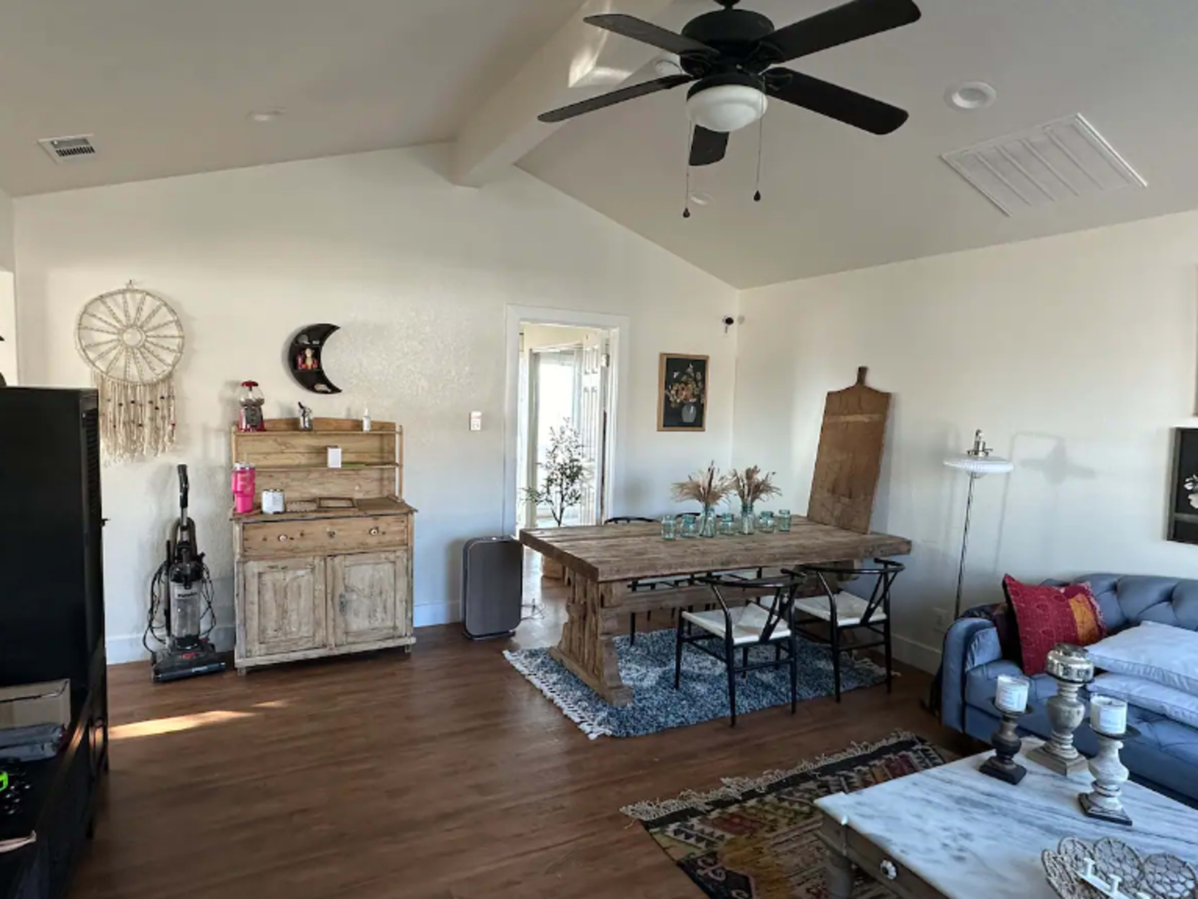 A well-lit living space featuring a wooden dining table with black chairs, a sofa, and decorative elements like a dreamcatcher and plants.