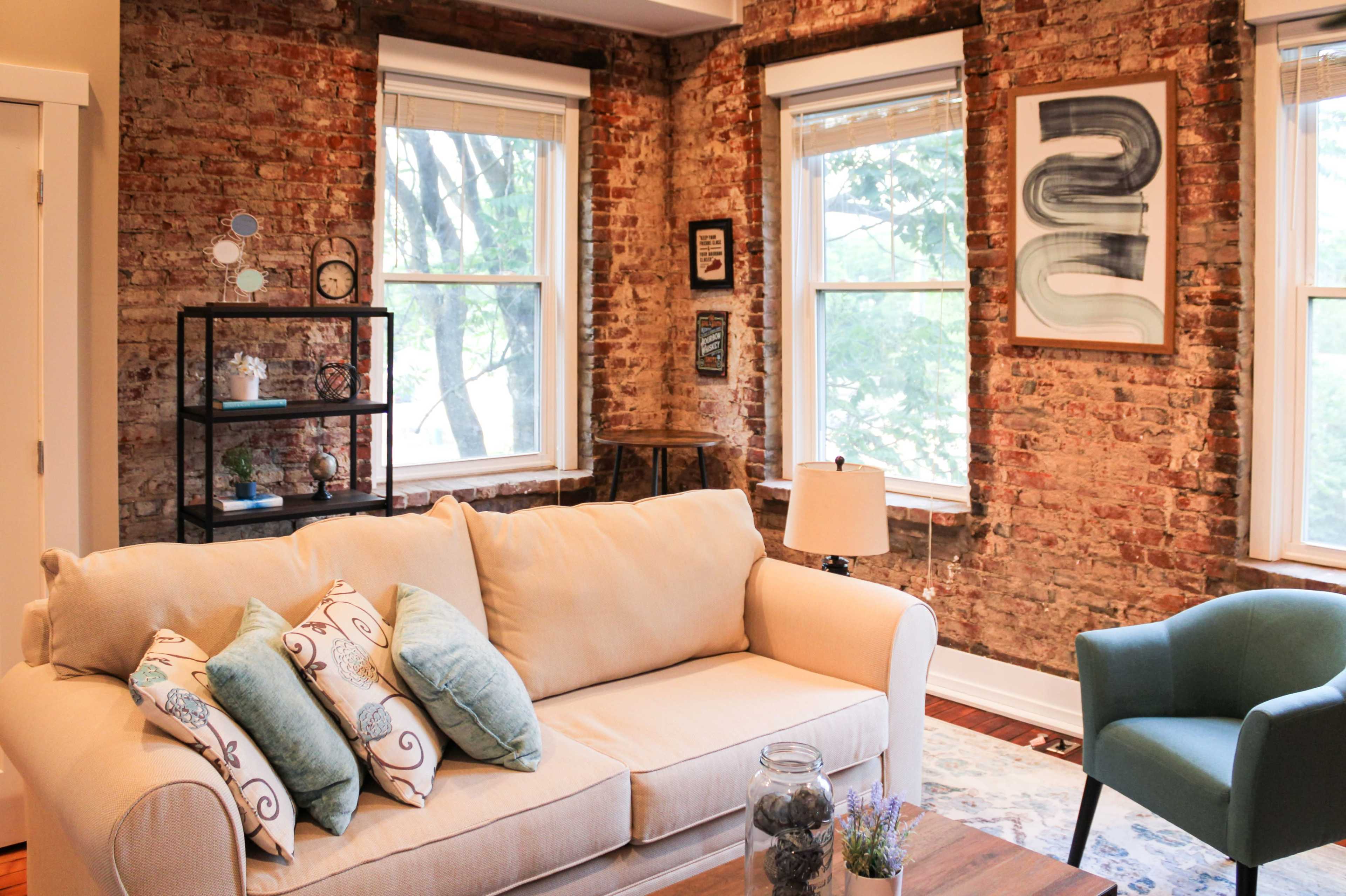 A cozy living room with a beige sofa adorned with decorative pillows, set against a backdrop of exposed brick walls and large windows.