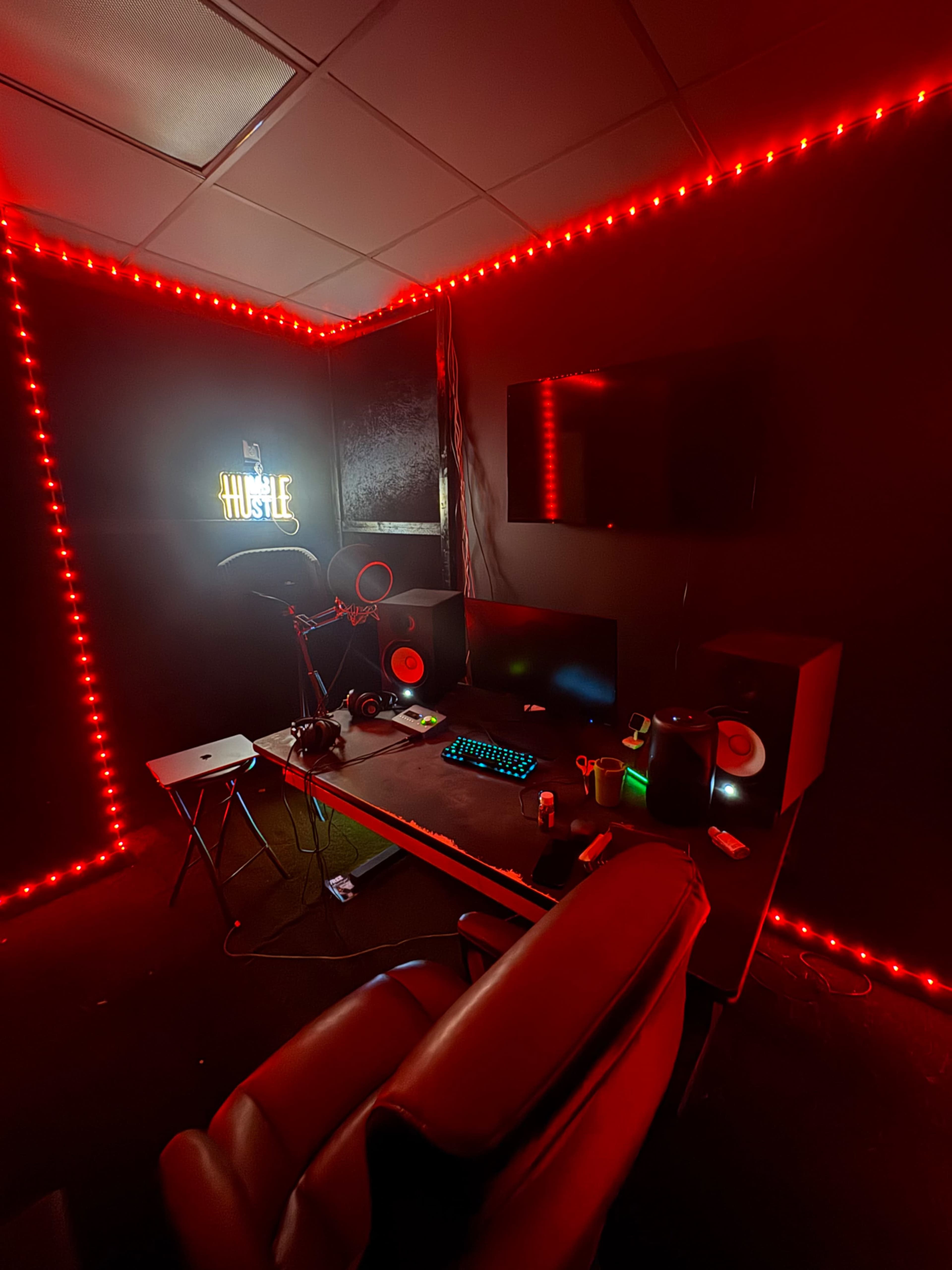 A dimly lit room features a desk with a computer setup, surrounded by red LED lights, a neon sign that reads "Hustle," and various audio equipment.