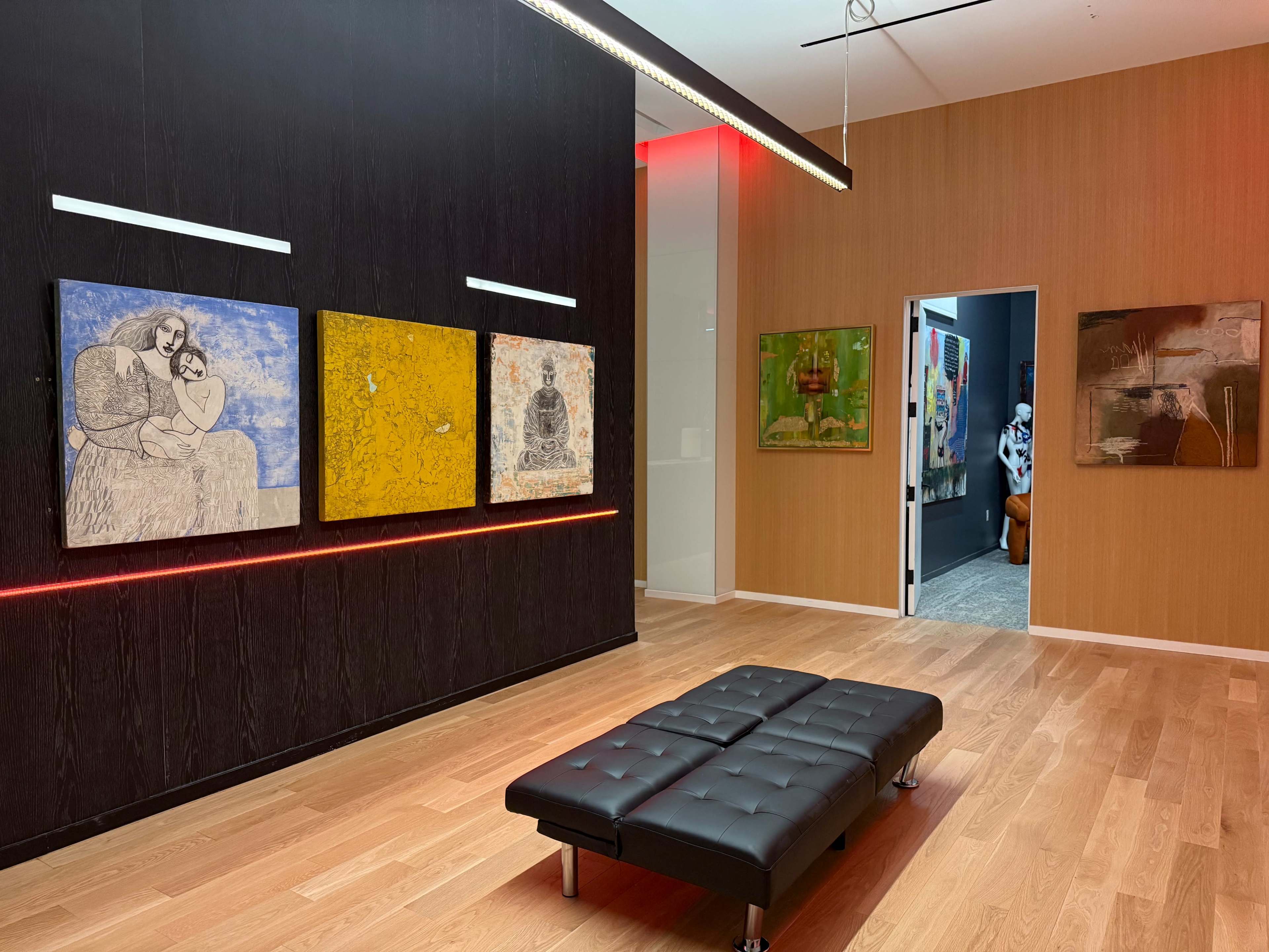 The image shows an art gallery featuring several paintings on the wall and a black leather bench in a warmly lit room with wooden flooring.