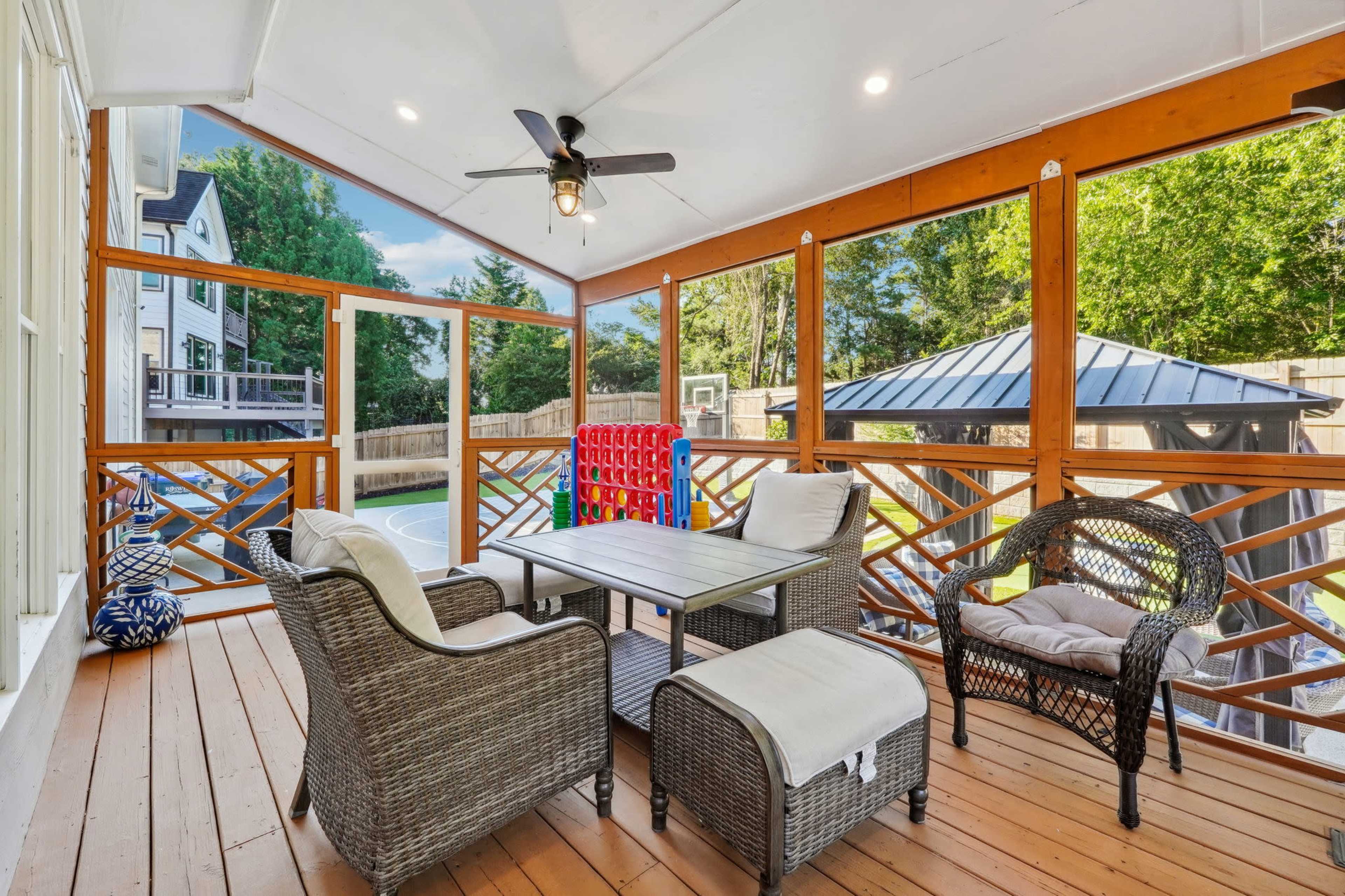 A screened porch with wicker furniture, a ceiling fan, and a view of a backyard featuring a basketball court and a gazebo.