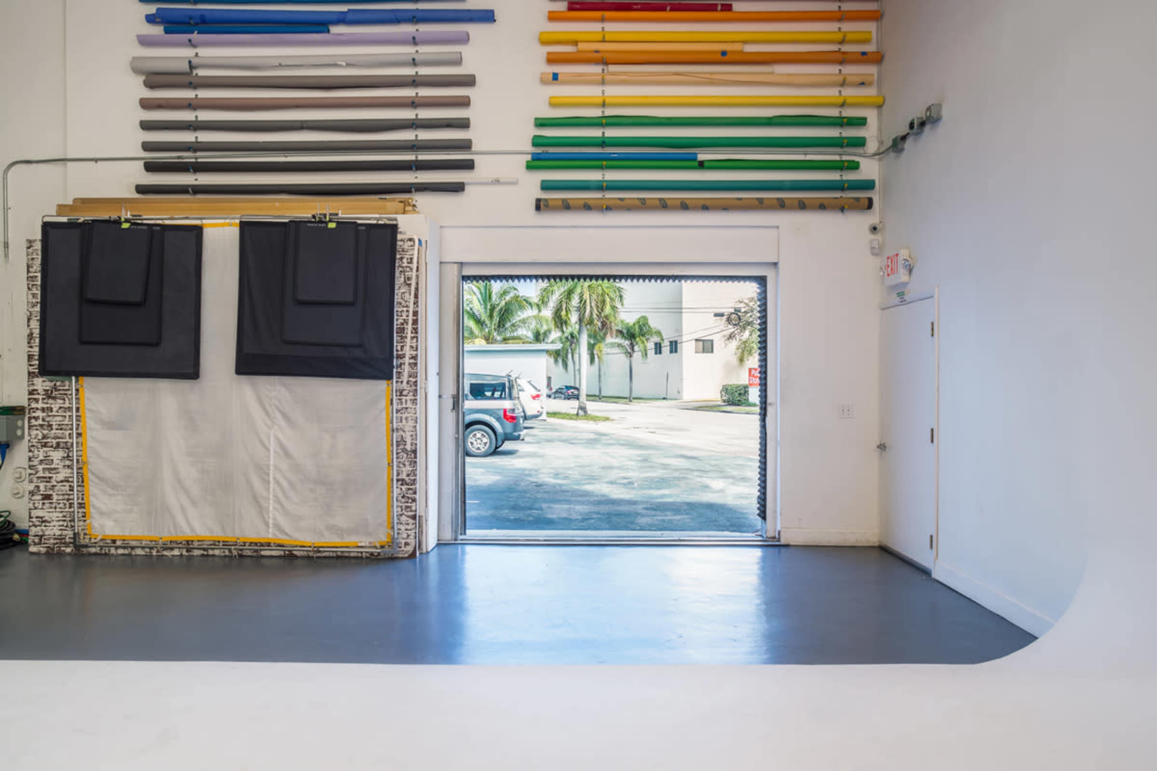 A warehouse interior with rolls of colored materials on the wall and an open door leading to a parking area outside.
