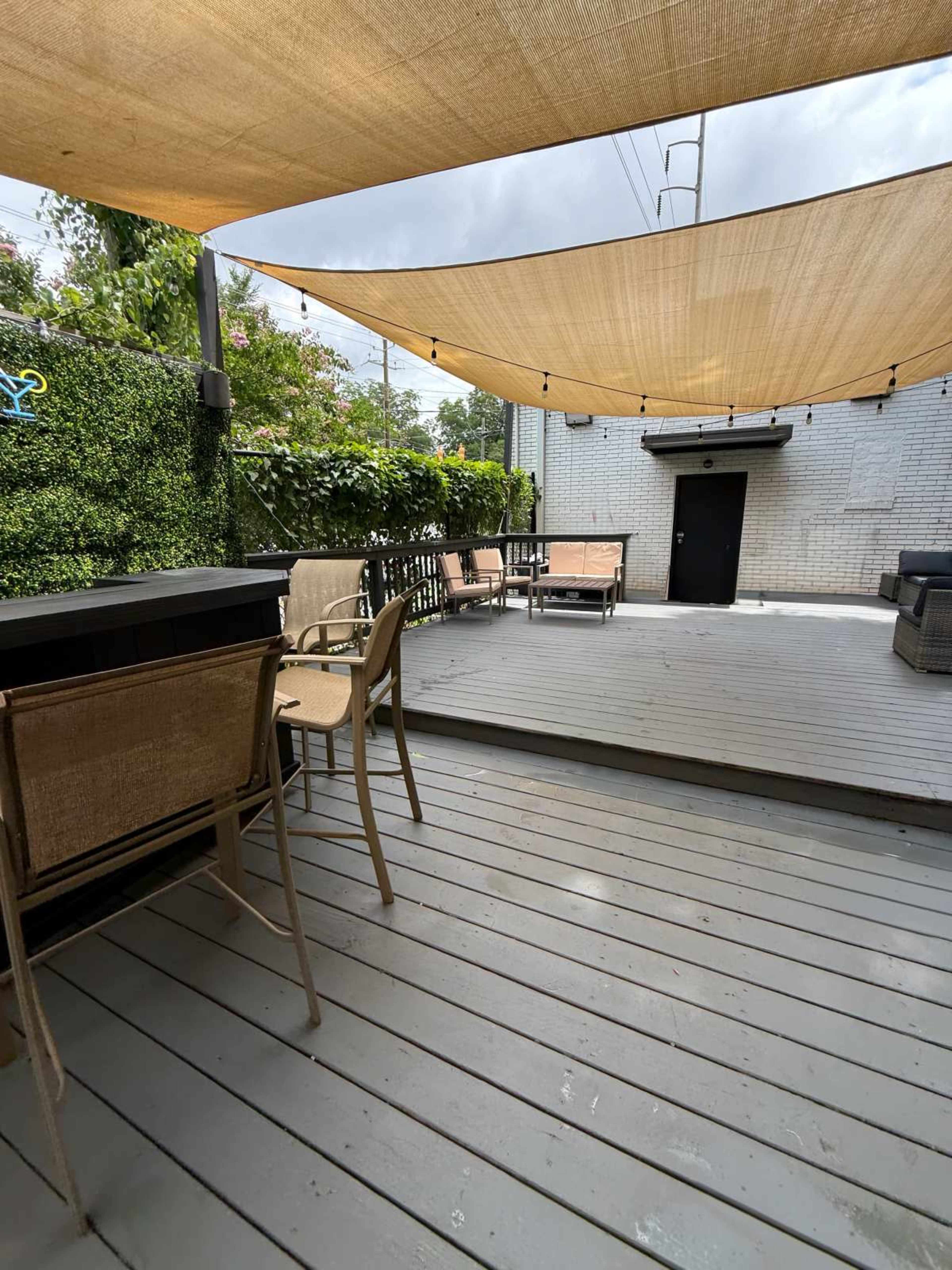 The image shows a spacious outdoor patio area with wooden flooring, shaded by large fabric canopies, and equipped with several chairs and a bar counter.