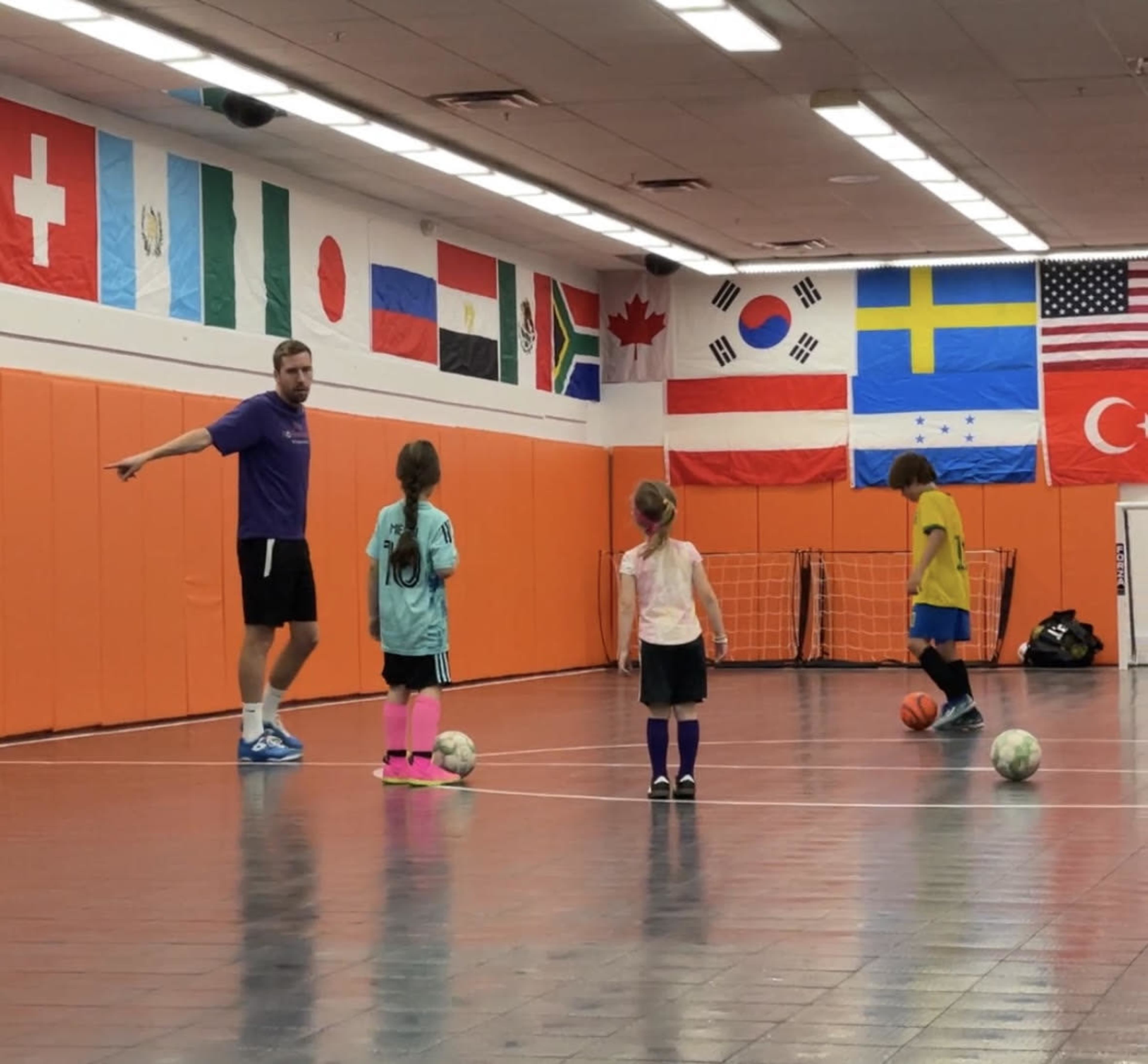 A coach directs a small group of children in a sports facility adorned with various national flags on the walls.