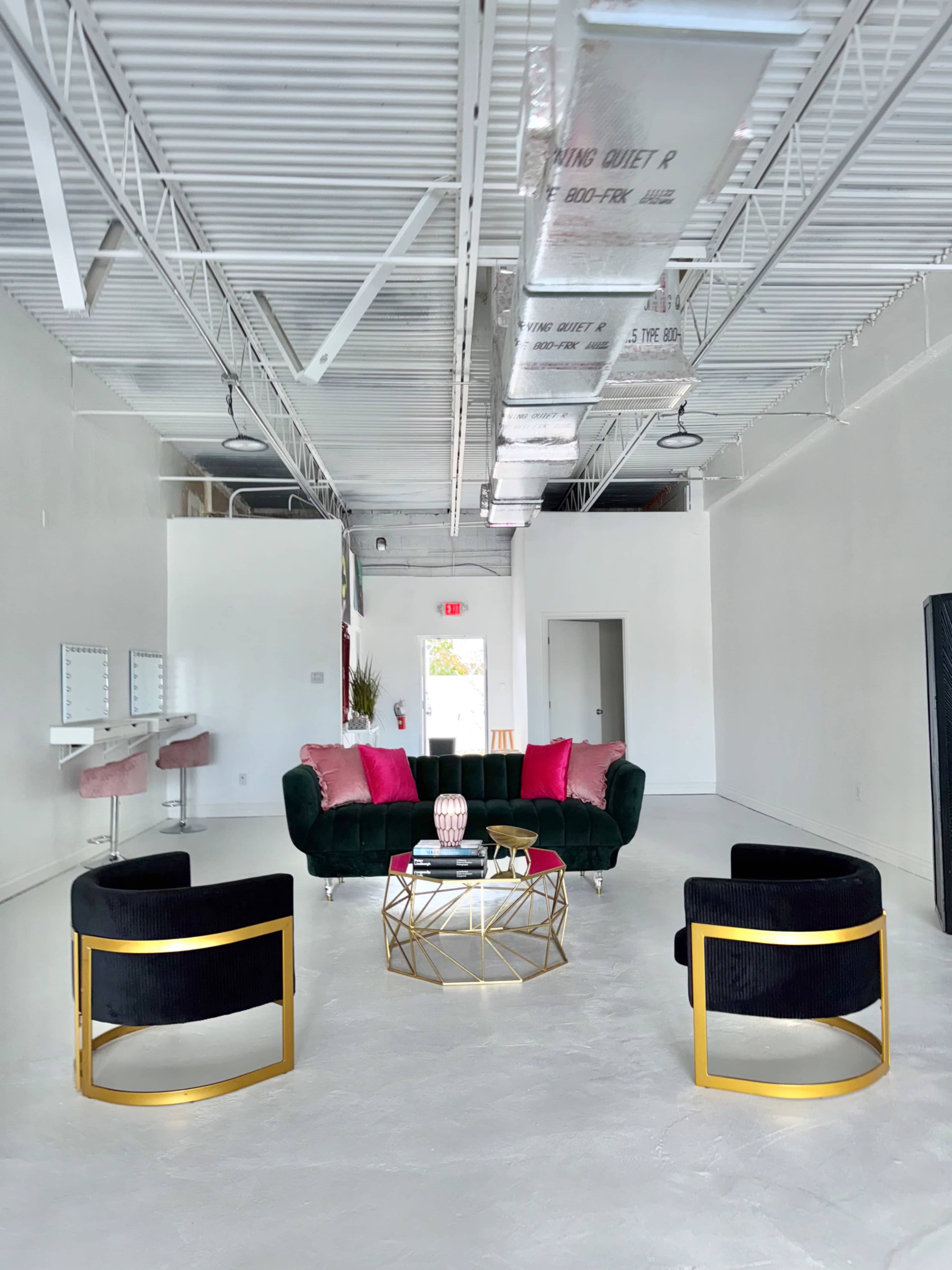 A spacious, modern room features a dark green sofa with pink accent pillows, two circular black chairs with gold frames, and a geometric coffee table.
