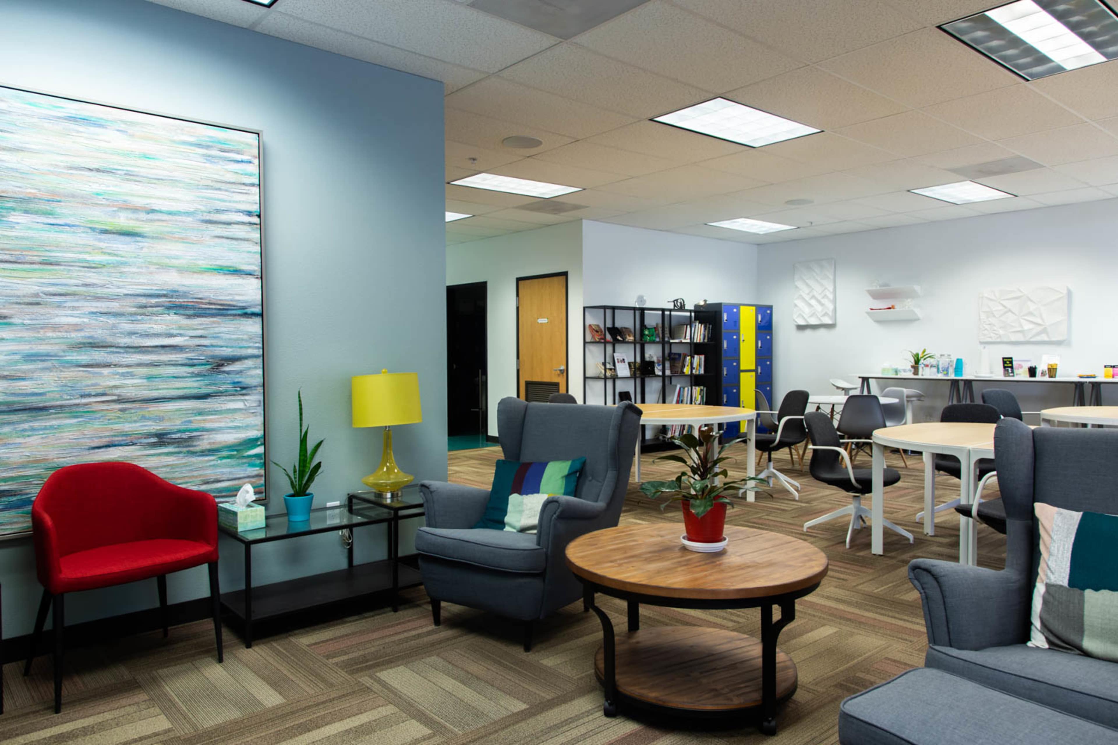 An office space featuring a seating area with chairs, a coffee table, and a decorative painting, alongside workstations equipped with desks and storage lockers.