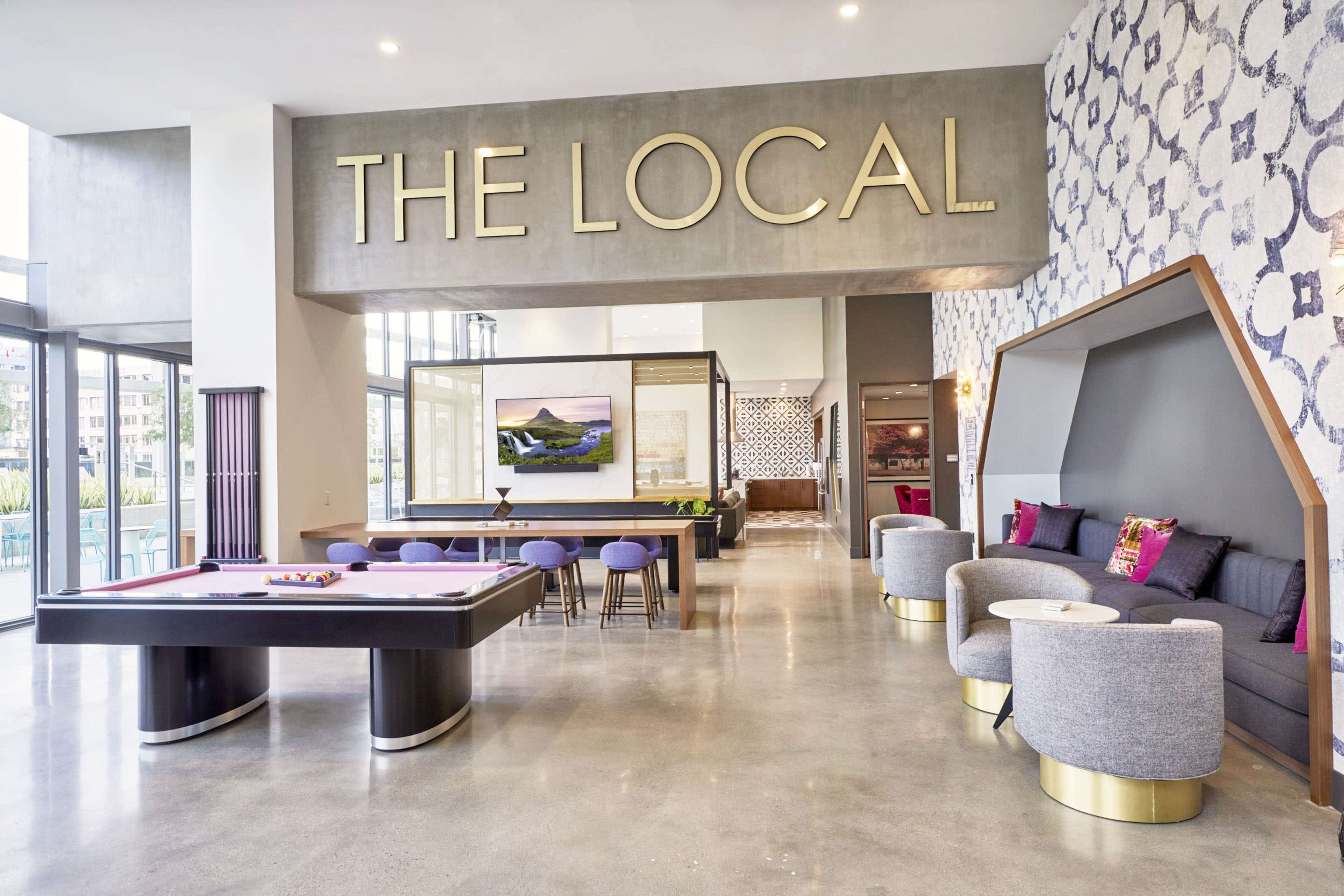 The image shows a modern lounge area with a pool table, seating arrangements, and large windows letting in natural light, featuring a prominent sign that reads "THE LOCAL."