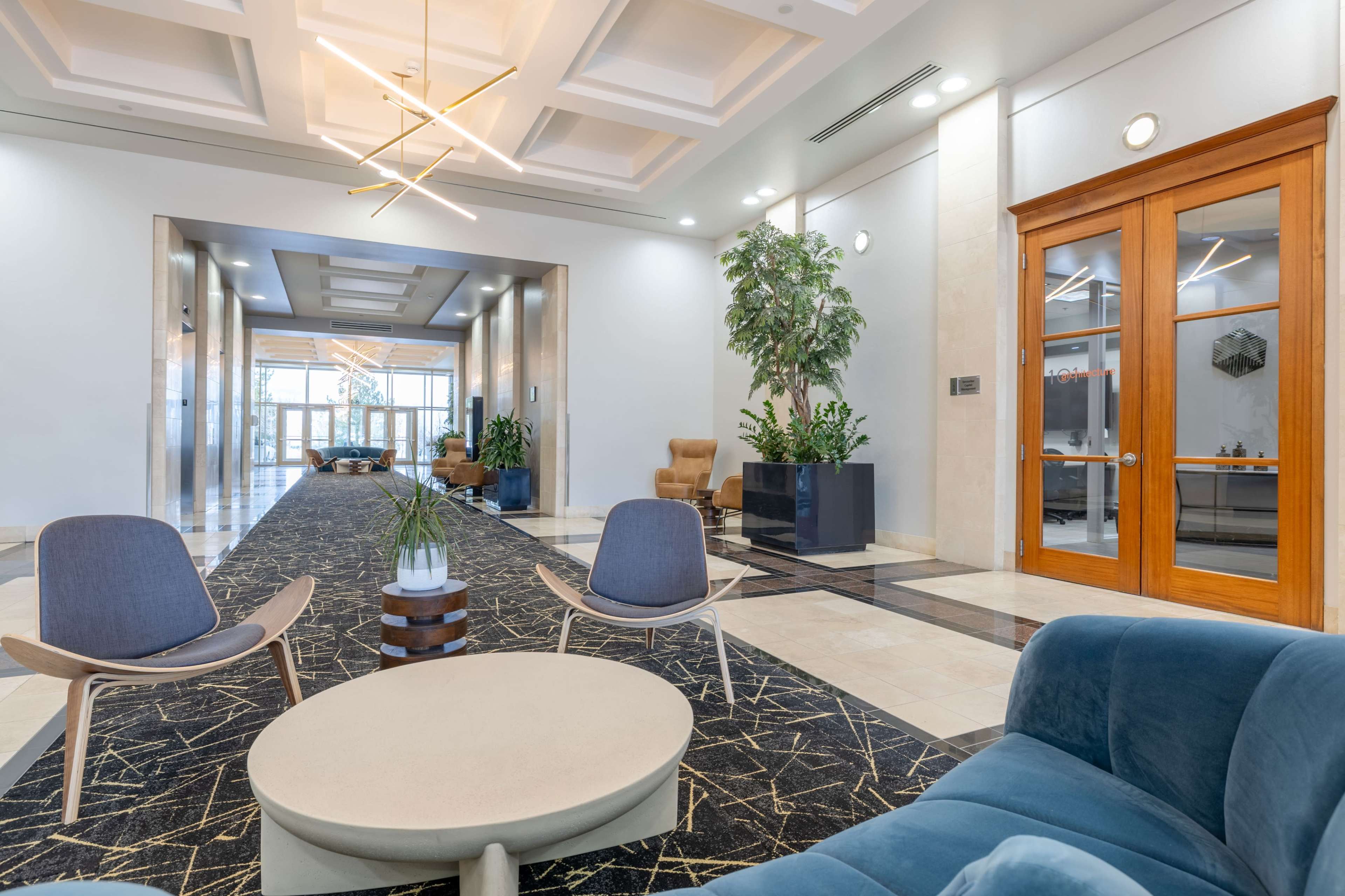 The image shows a modern hotel lobby featuring two lounge chairs, a round coffee table, and decorative plants, with glass doors leading to a corridor.