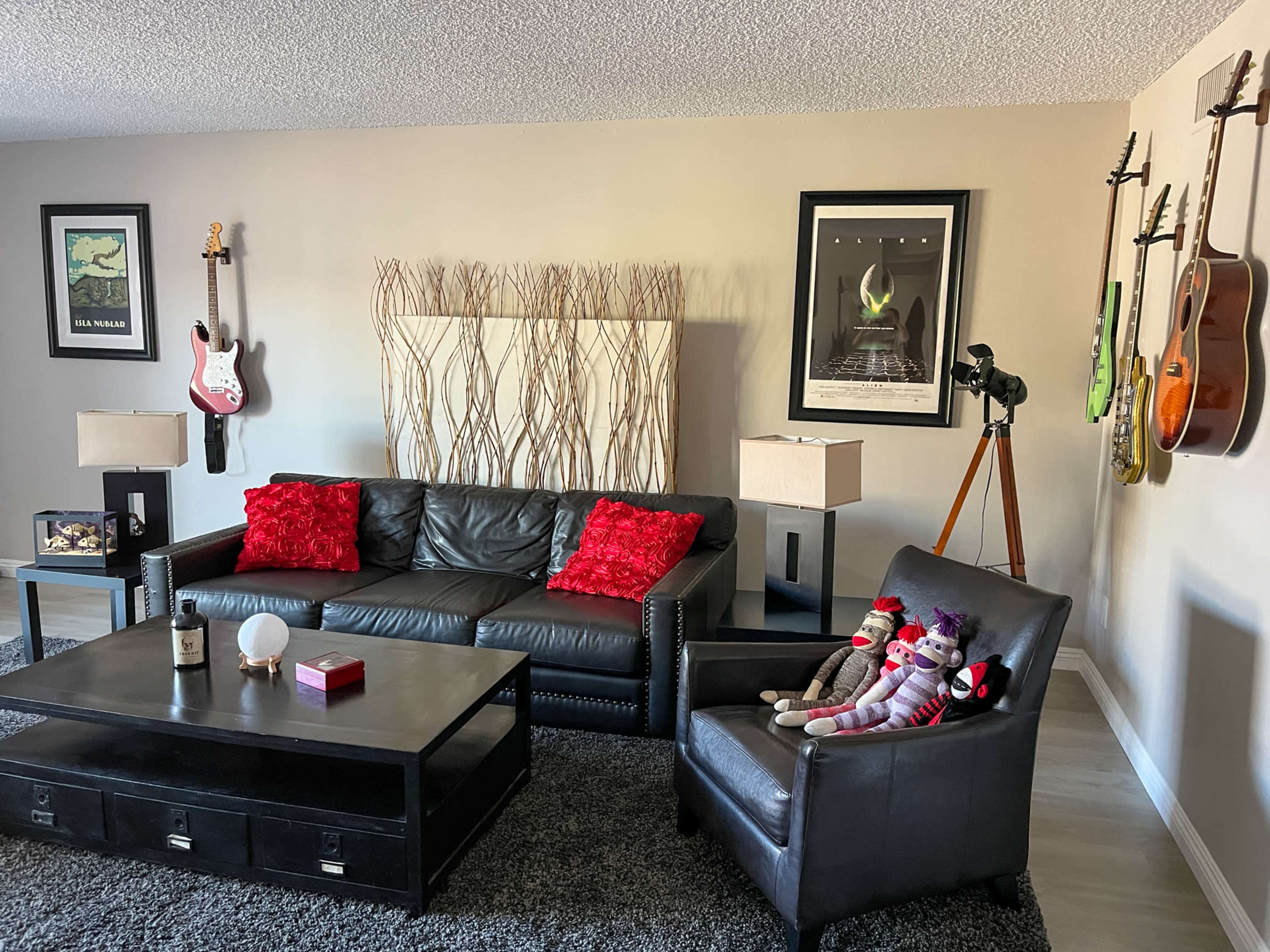 The living room features a black leather couch with red cushions, a coffee table, two lamps, and several musical instruments displayed on the walls.