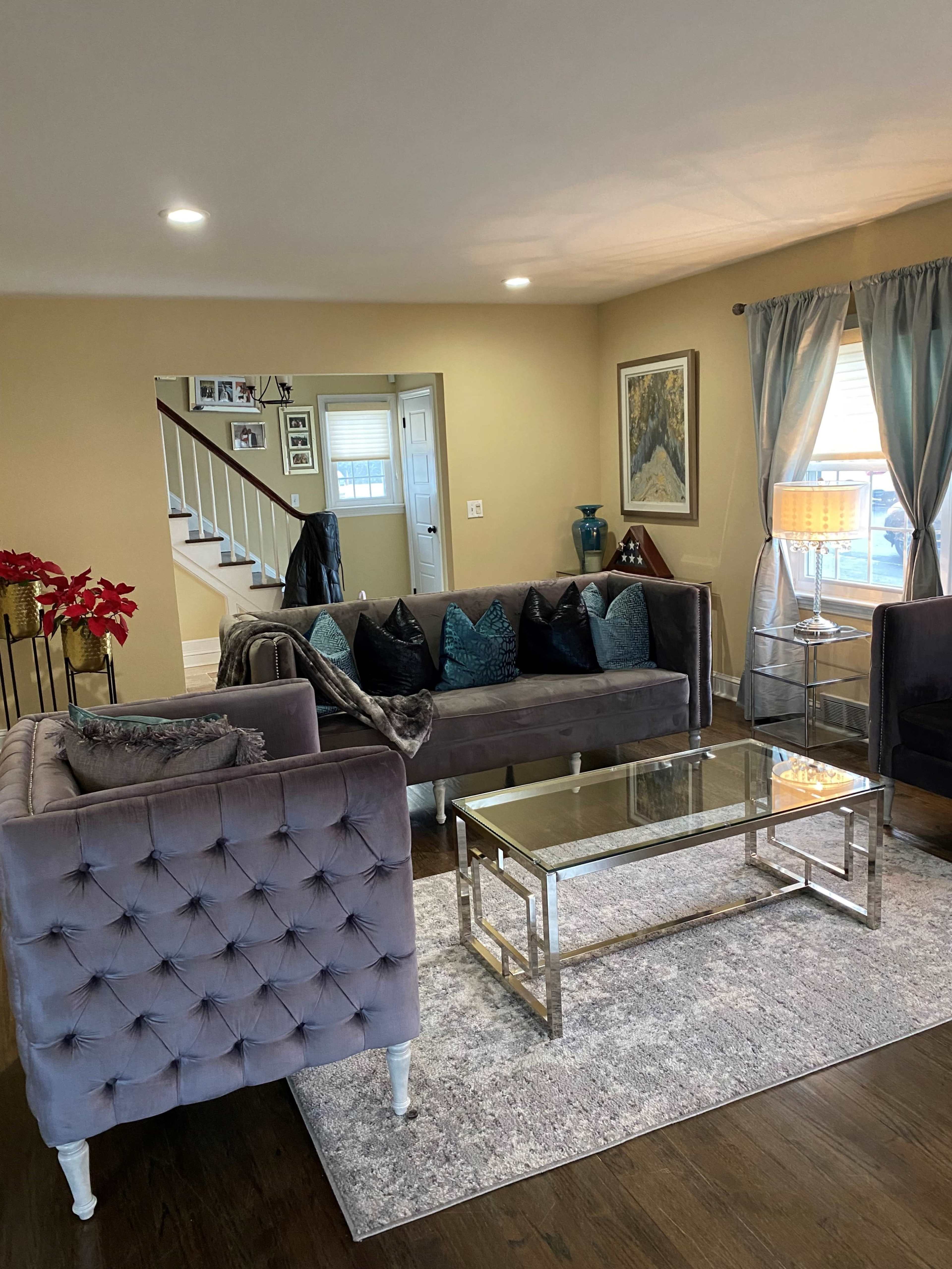 The living room features two velvet sofas, a glass coffee table, and decorative pillows arranged on the seating, with a light-colored wall and a staircase visible in the background.