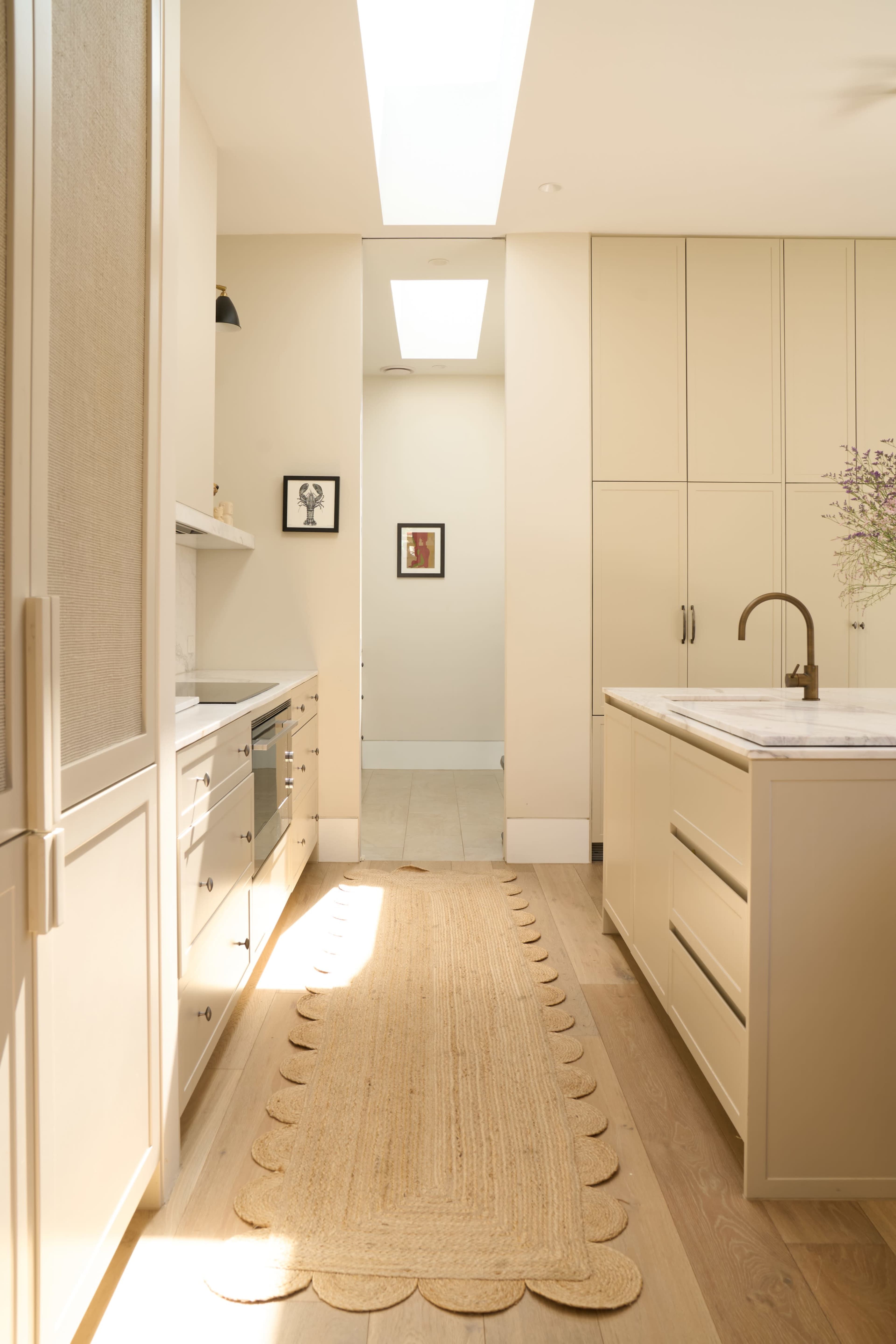 A kitchen with light-colored cabinets, a central island, and a jute runner leading to a doorway illuminated by a skylight.