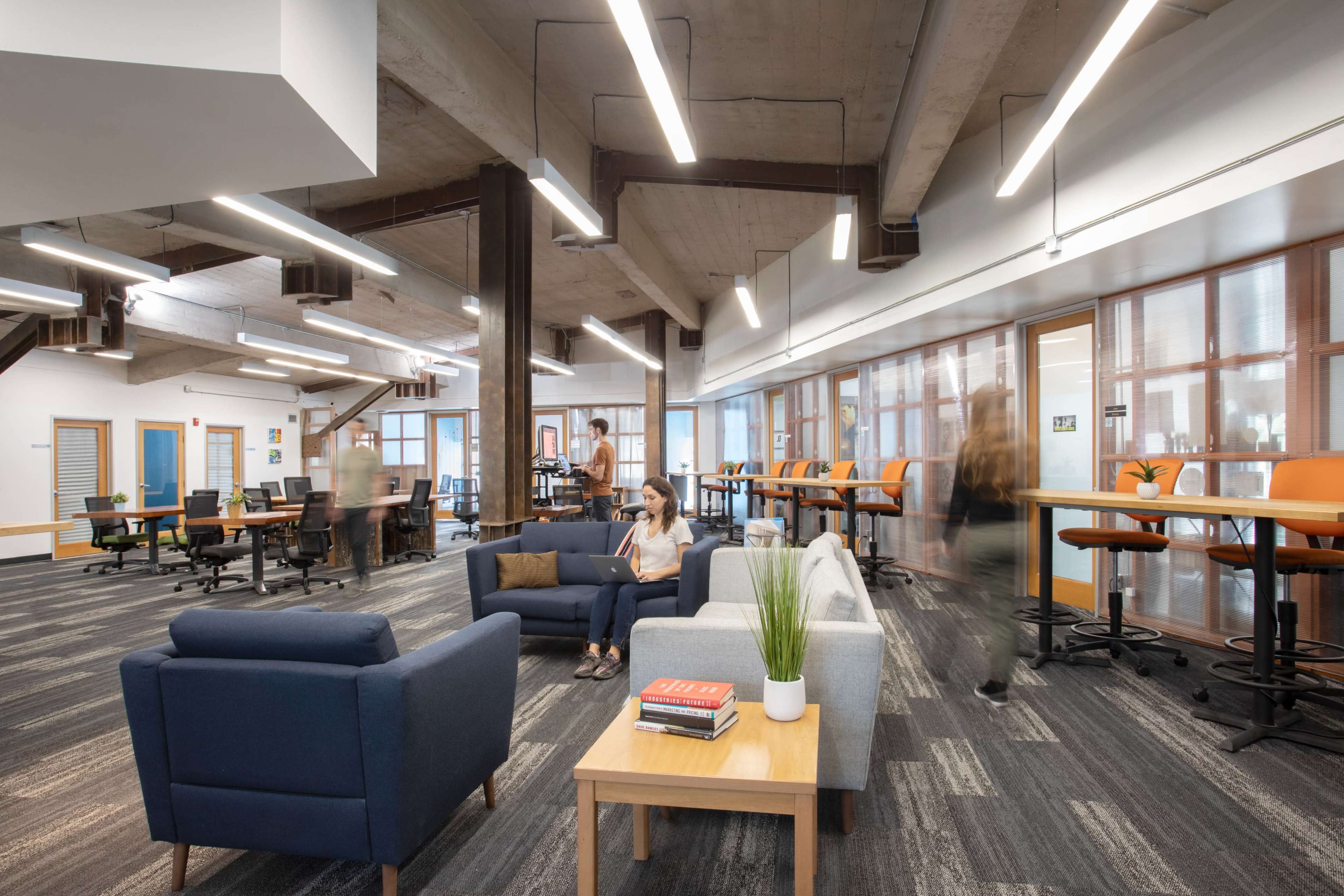 A modern office space features a seating area with sofas, a coffee table, and multiple workstations in the background.