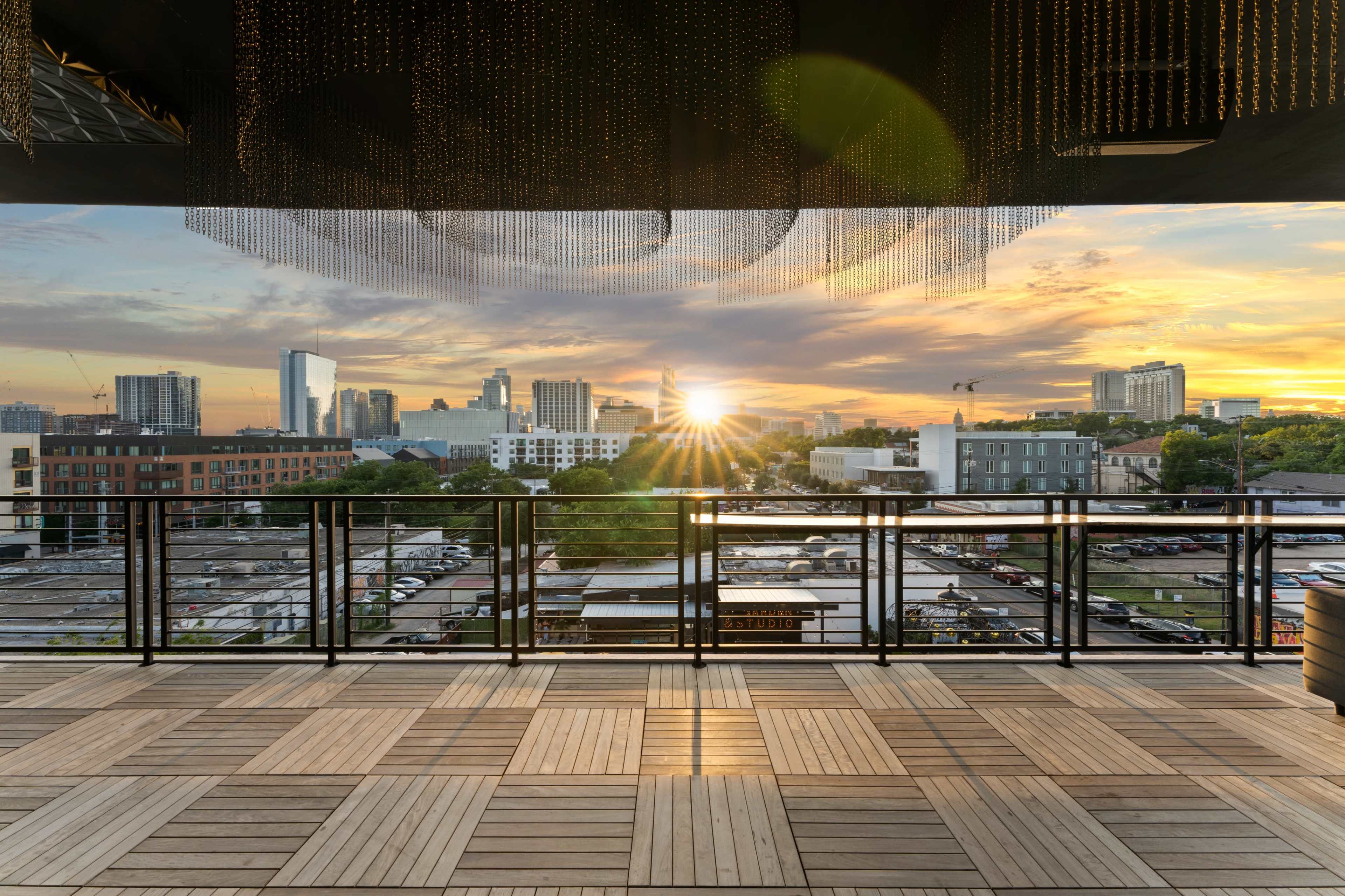A rooftop terrace with wooden flooring overlooking a city skyline at sunset, featuring buildings and a radiant sun in the background.