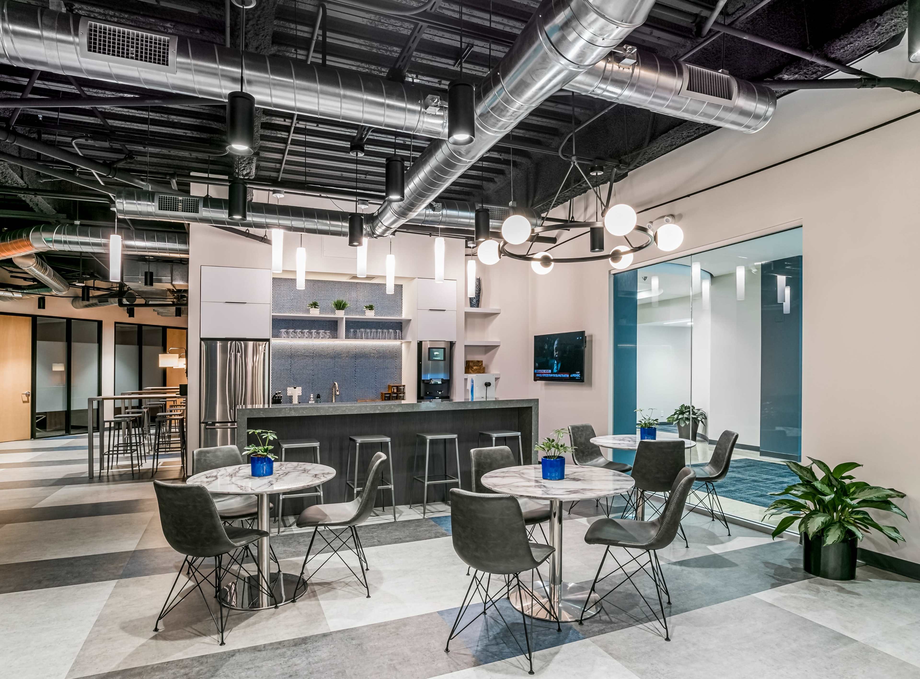 A modern office kitchen area with a bar, tables, and chairs, featuring industrial-style light fixtures and plants.