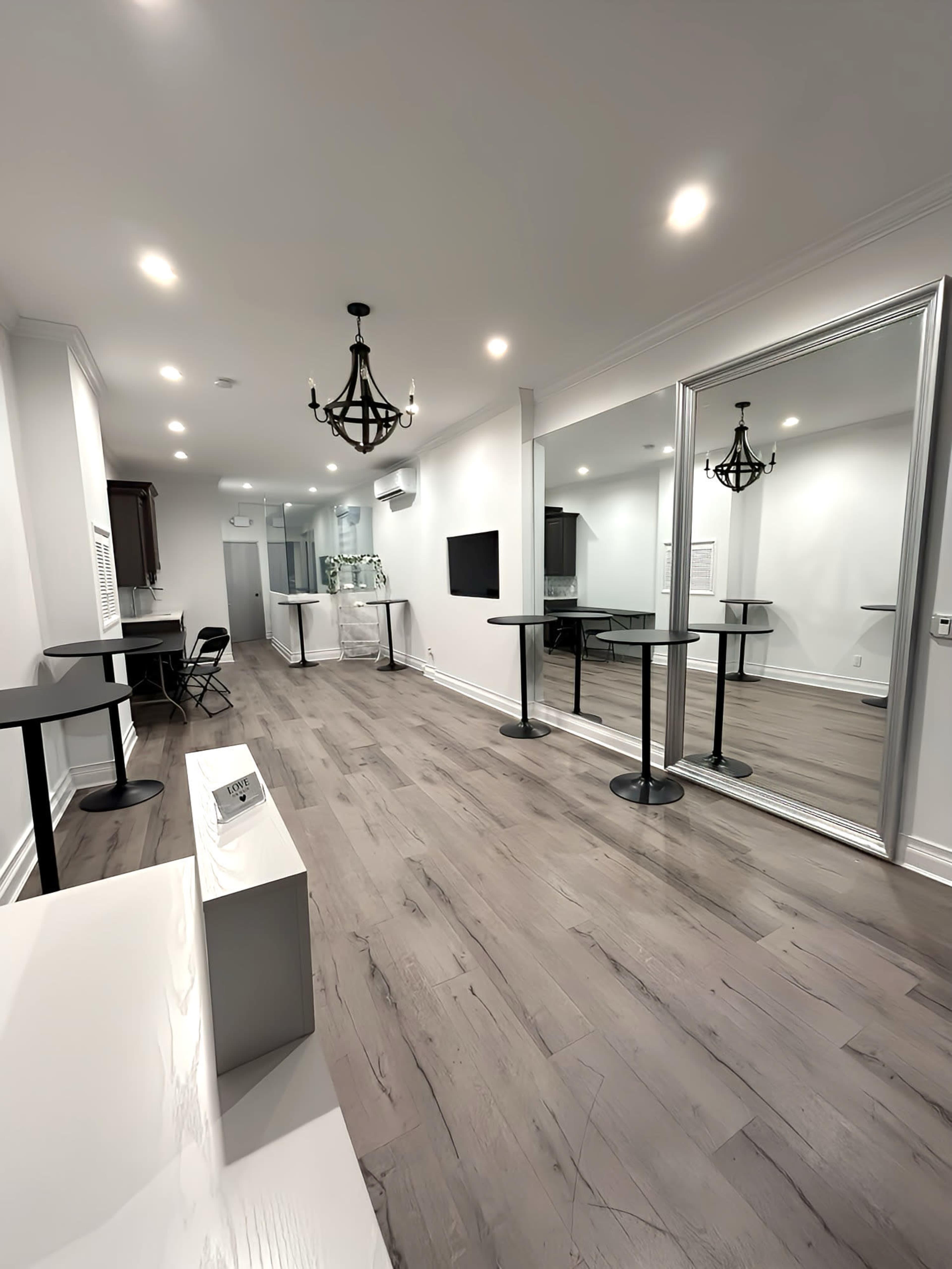 The image shows a modern interior space with light wood flooring, a large wall mirror, and tables arranged for use, illuminated by overhead lighting and a chandelier.