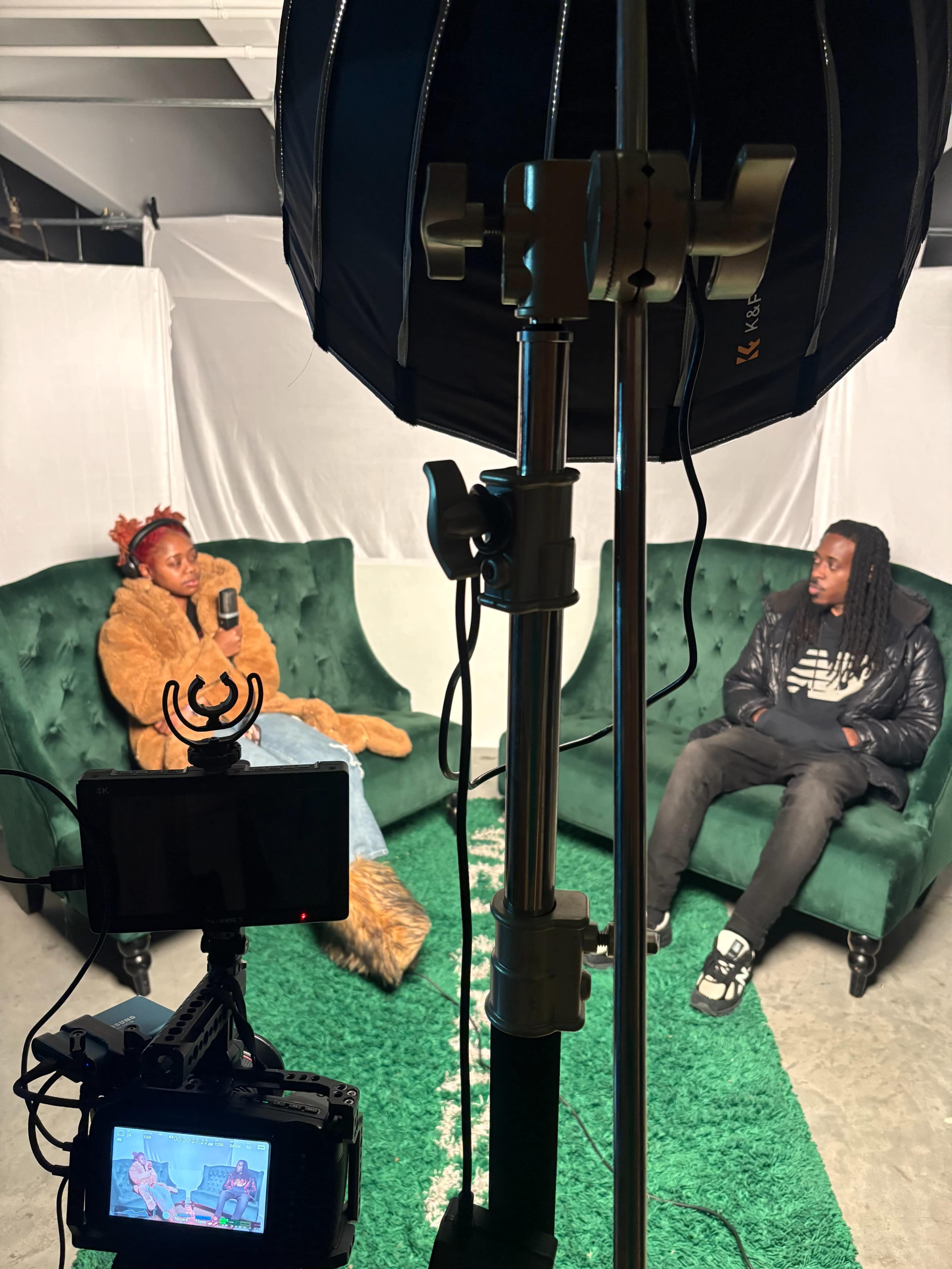 The image shows two individuals seated on green couches in a studio setting, with a camera and lighting equipment positioned in the foreground.