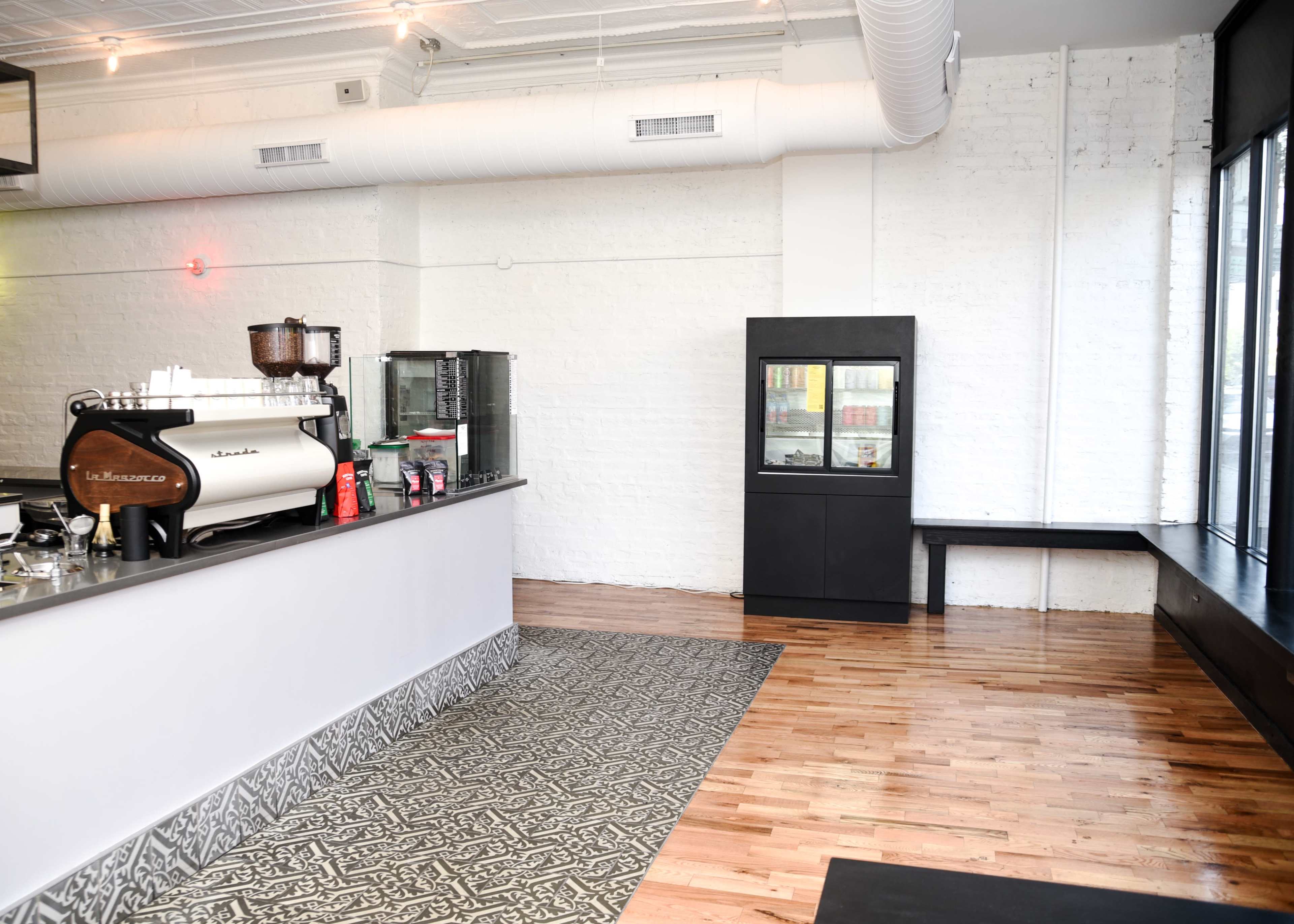 A minimalist café interior with a counter, coffee machine, and a glass display refrigerator, set against a white brick wall and wooden flooring.
