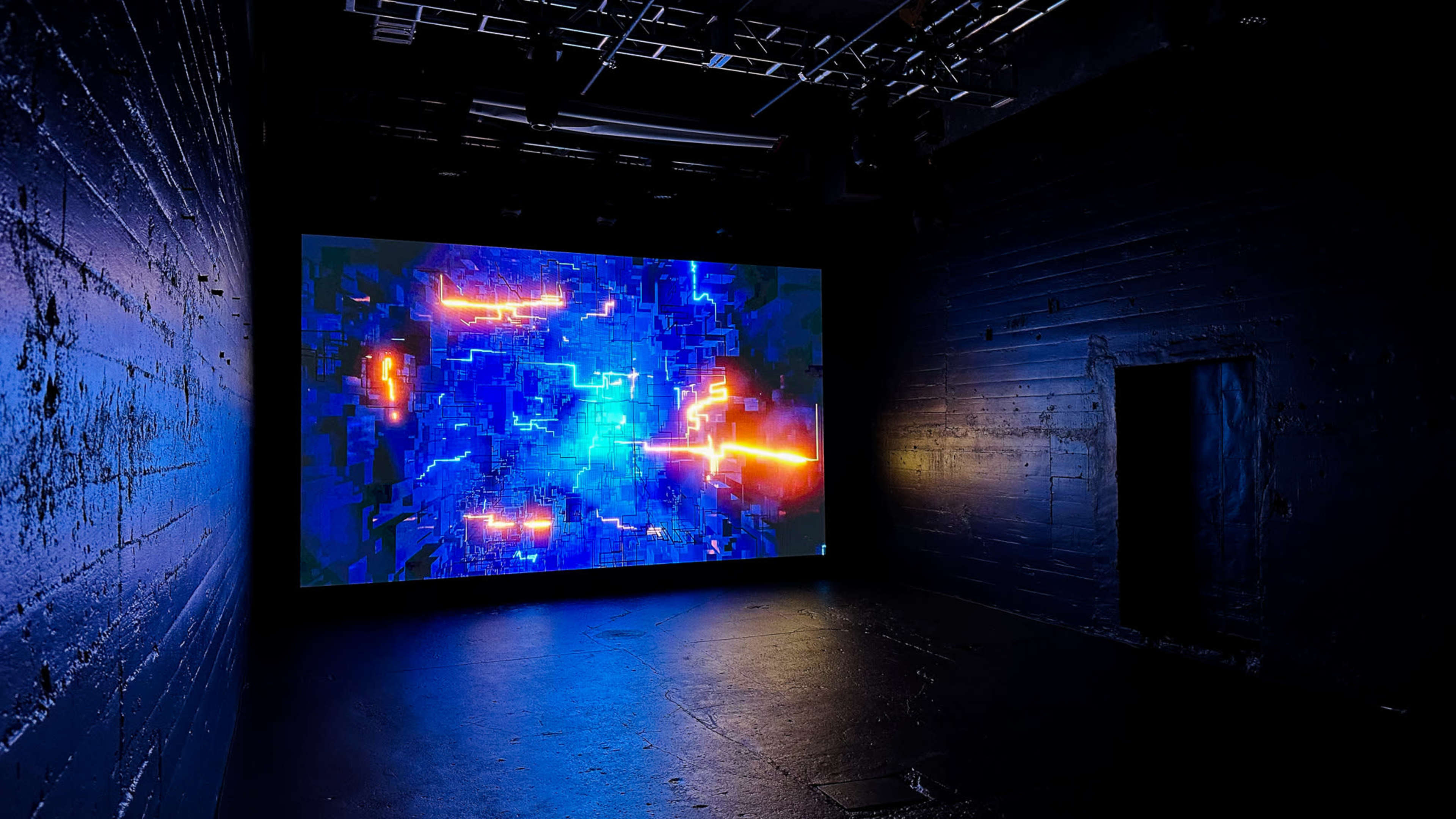 A large screen displays a vibrant abstract digital artwork in a dimly lit room with dark walls.