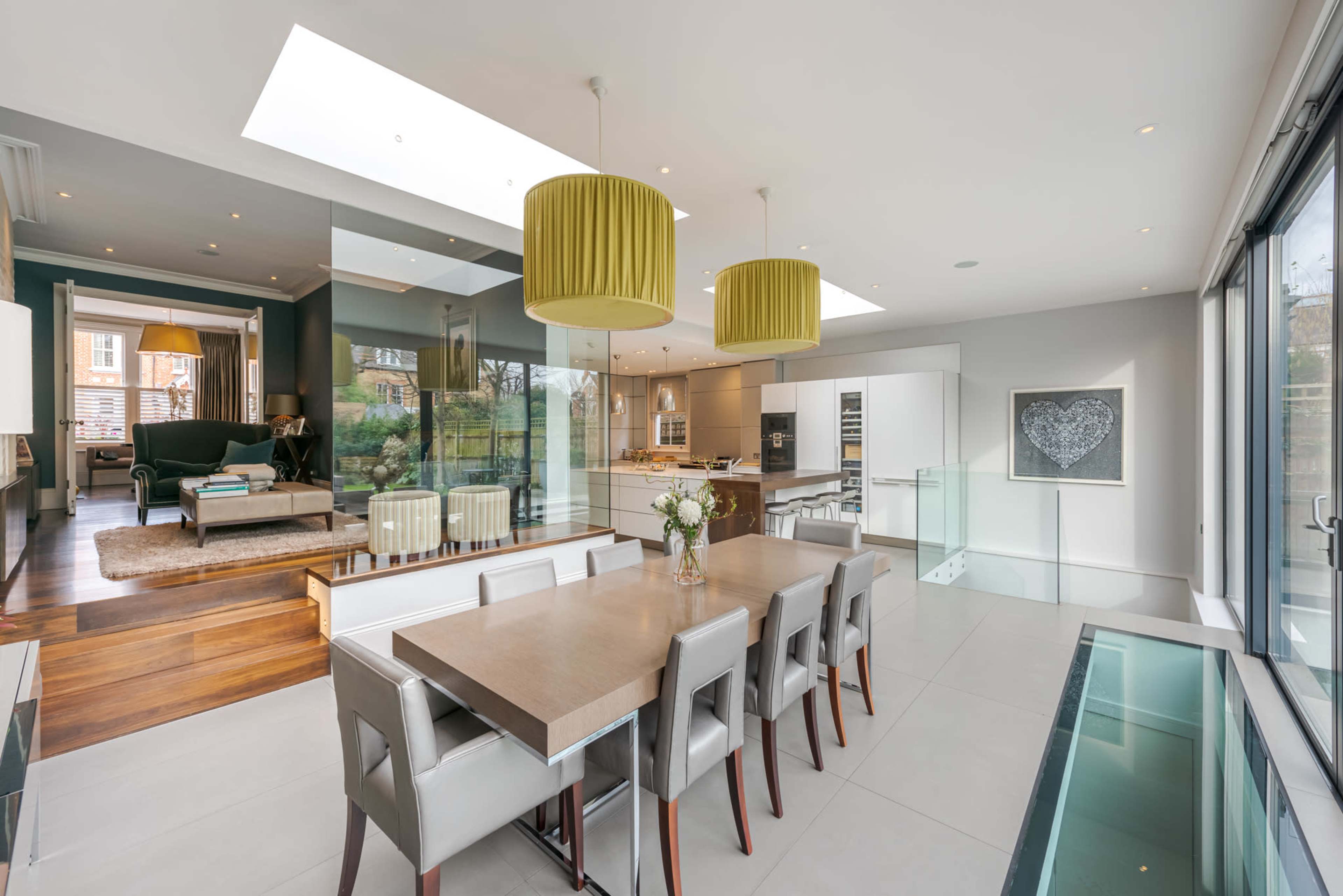 A modern, open-concept kitchen and dining area features a long wooden table surrounded by gray chairs, large windows providing natural light, and contemporary pendant lights.