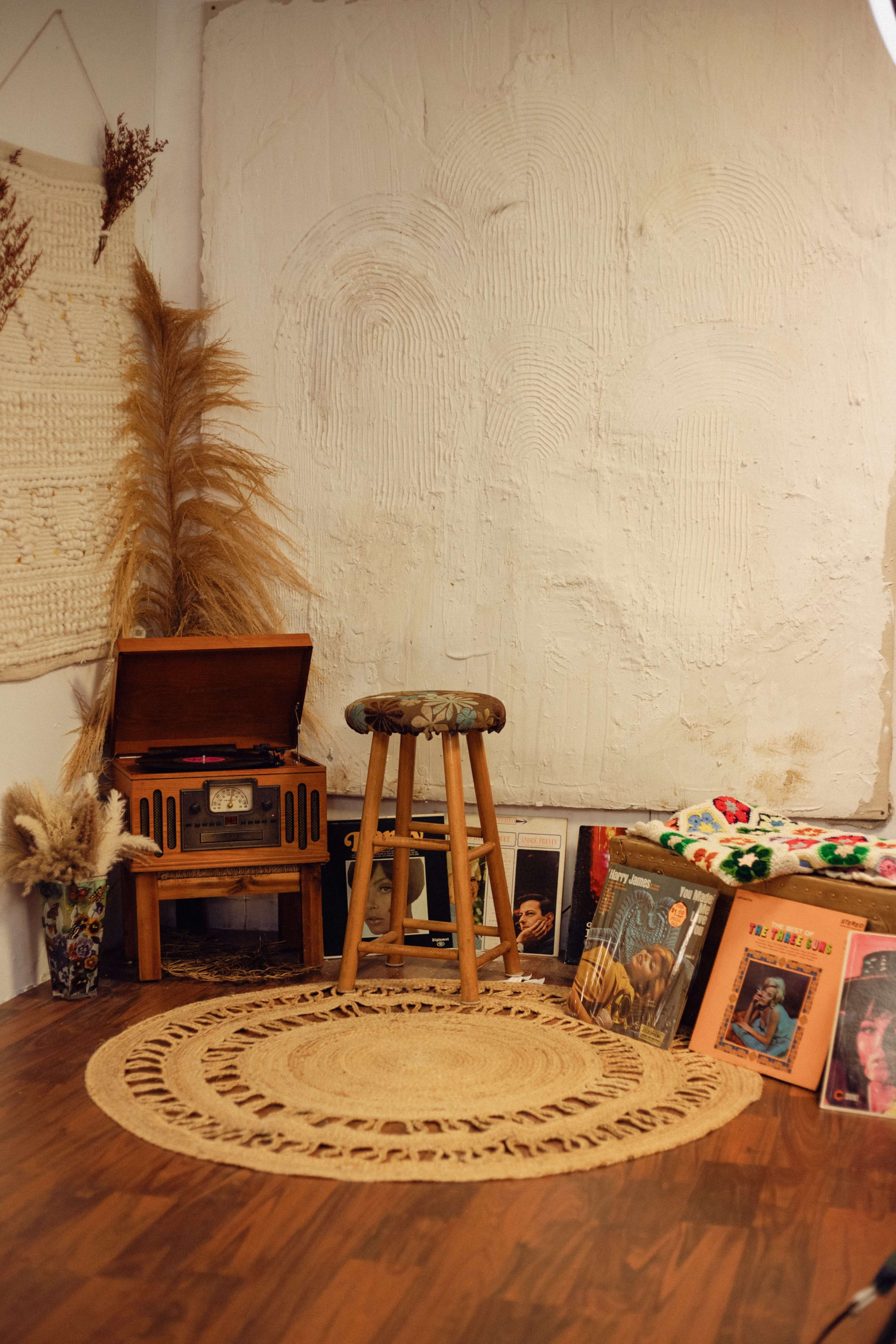 A corner of a room features a wooden record player, a bar stool, a round woven rug, and a collection of vinyl records displayed nearby.