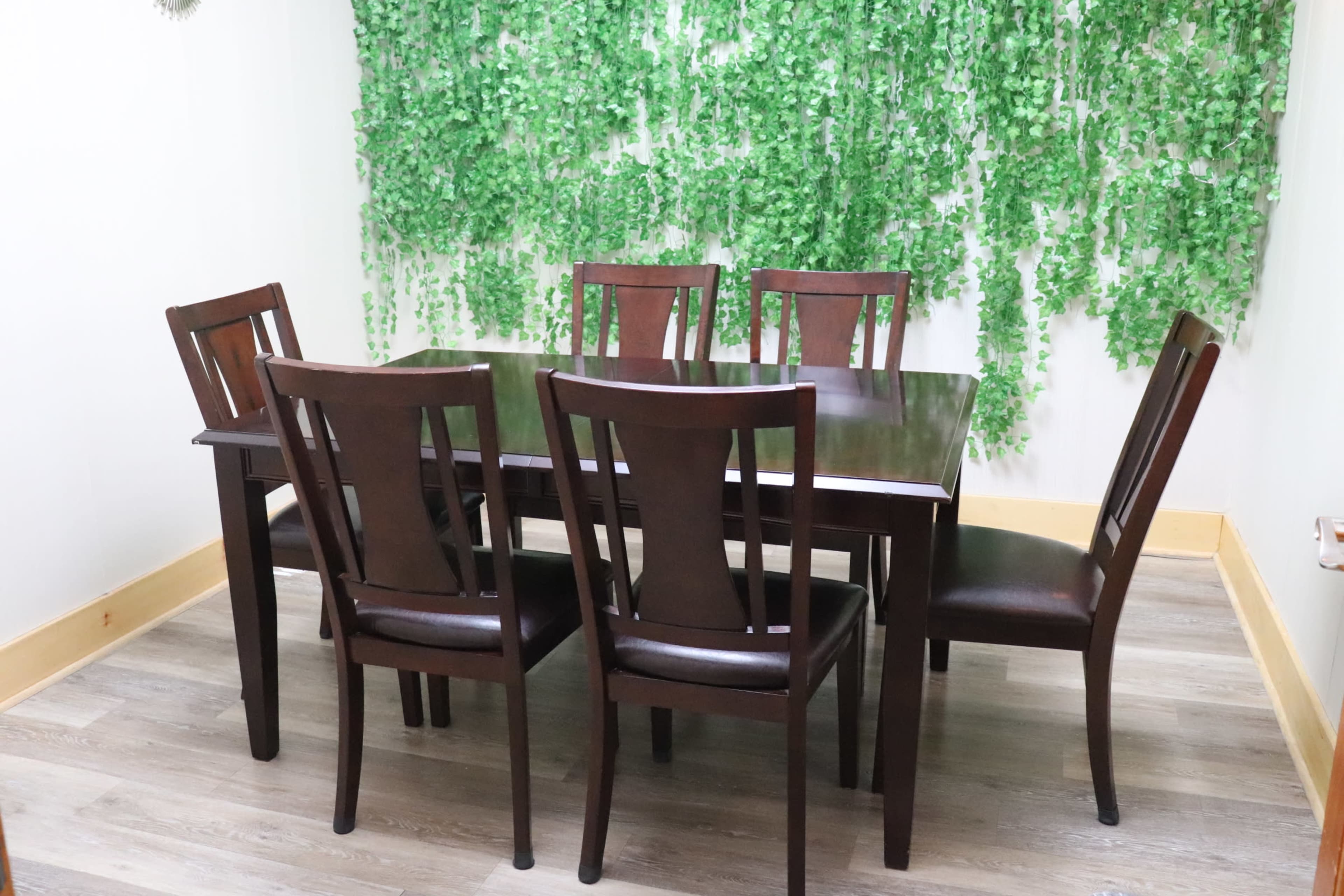 A wooden dining table surrounded by six matching chairs is set against a backdrop of green foliage.