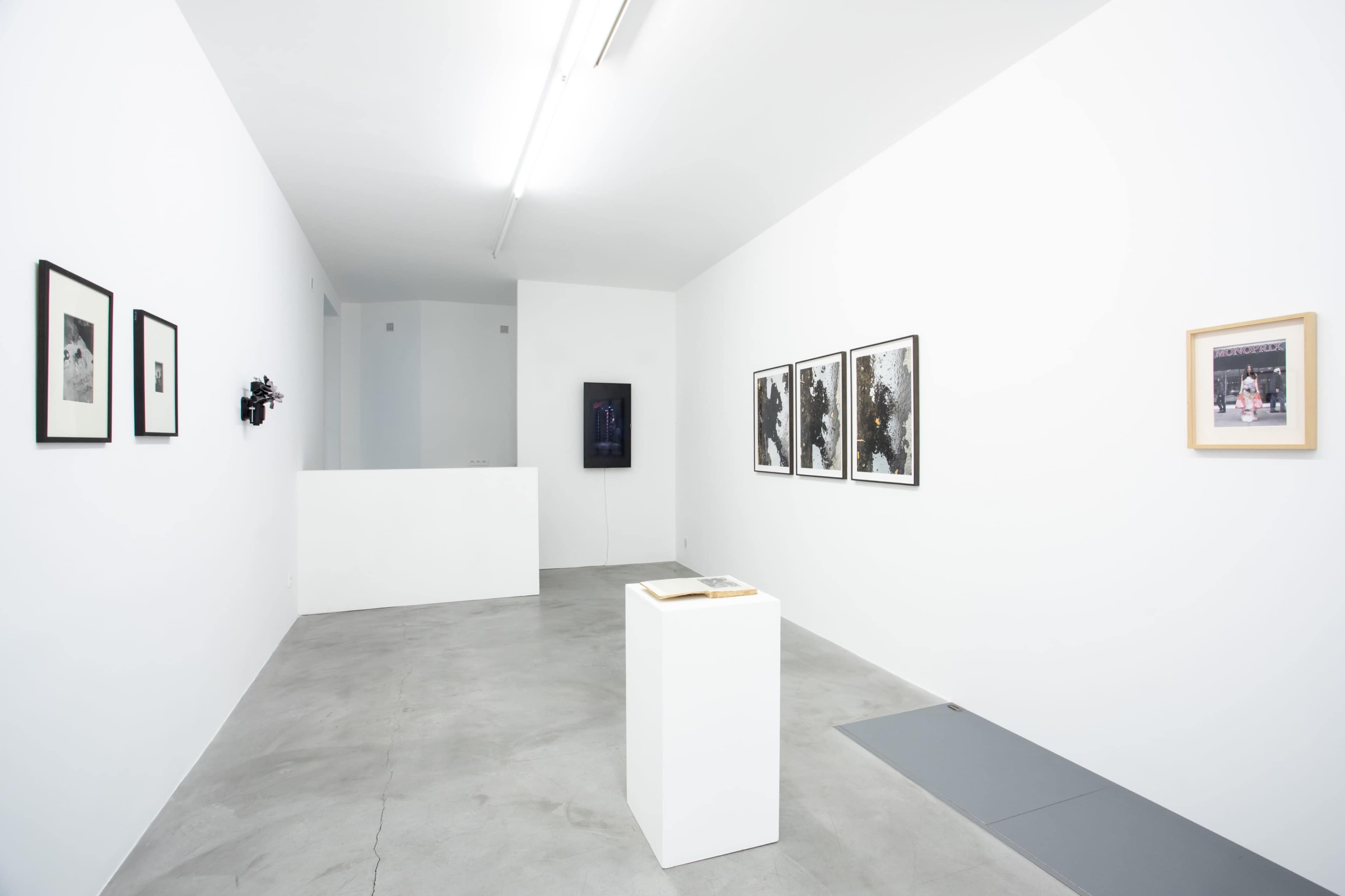 The image shows a minimalist art gallery with white walls, concrete flooring, and several framed artworks displayed on the walls, accompanied by a pedestal in the center.