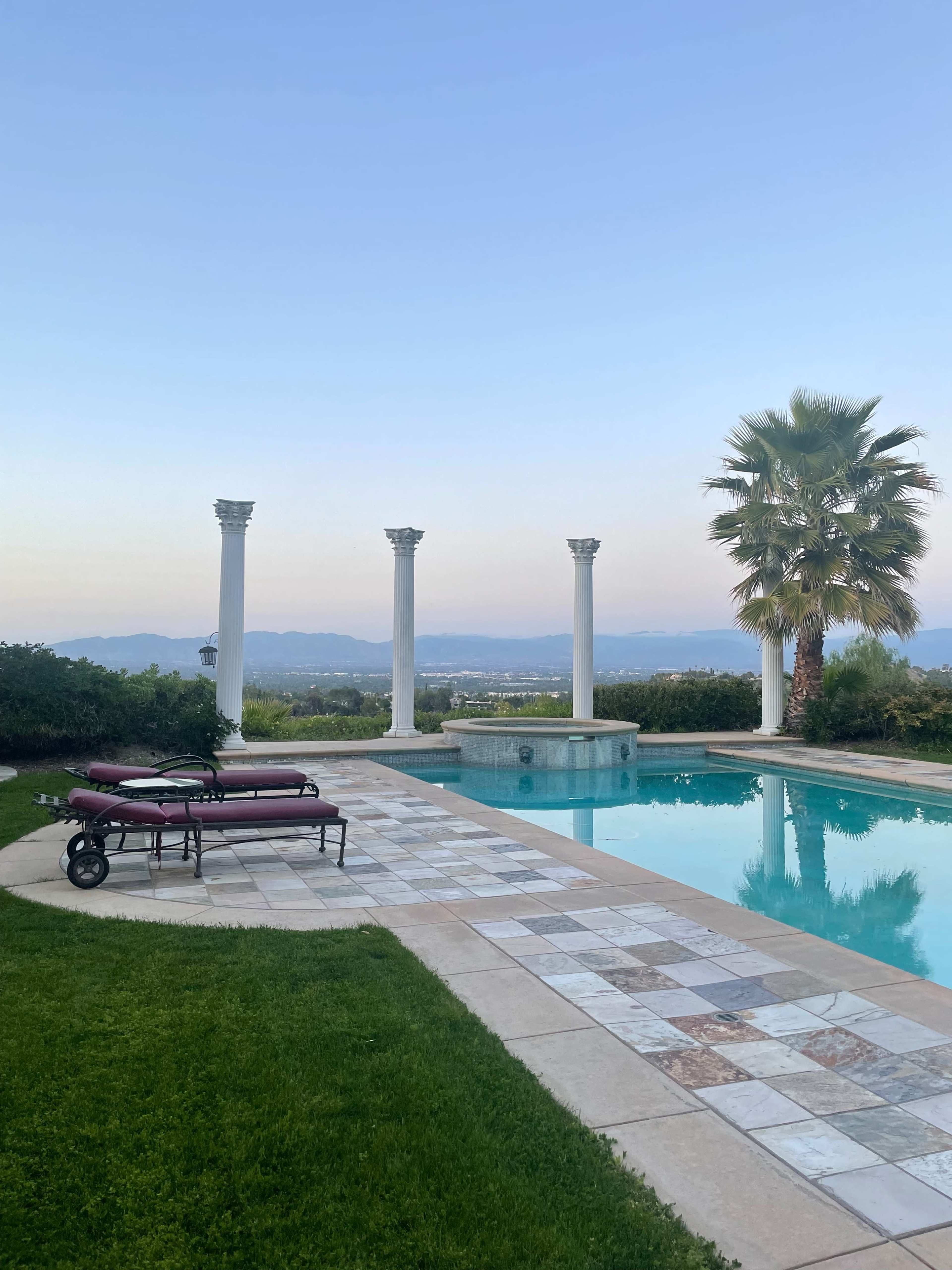 A tranquil pool area with marble tiles, a palm tree, and four white columns overlooking a scenic landscape.