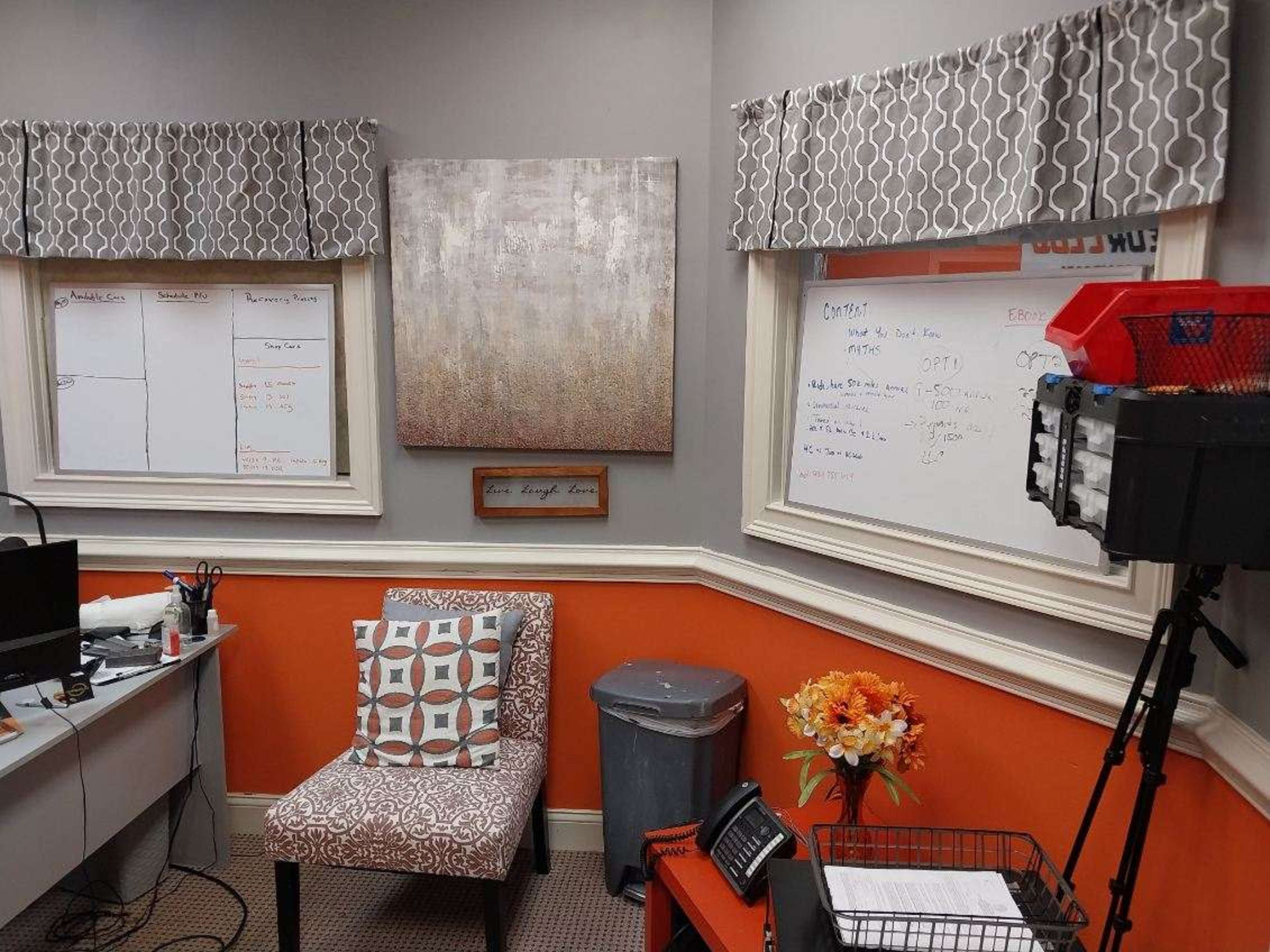 A small office with a gray chair, a whiteboard, and decorative curtains above two windows, featuring an orange wall and a trash can.