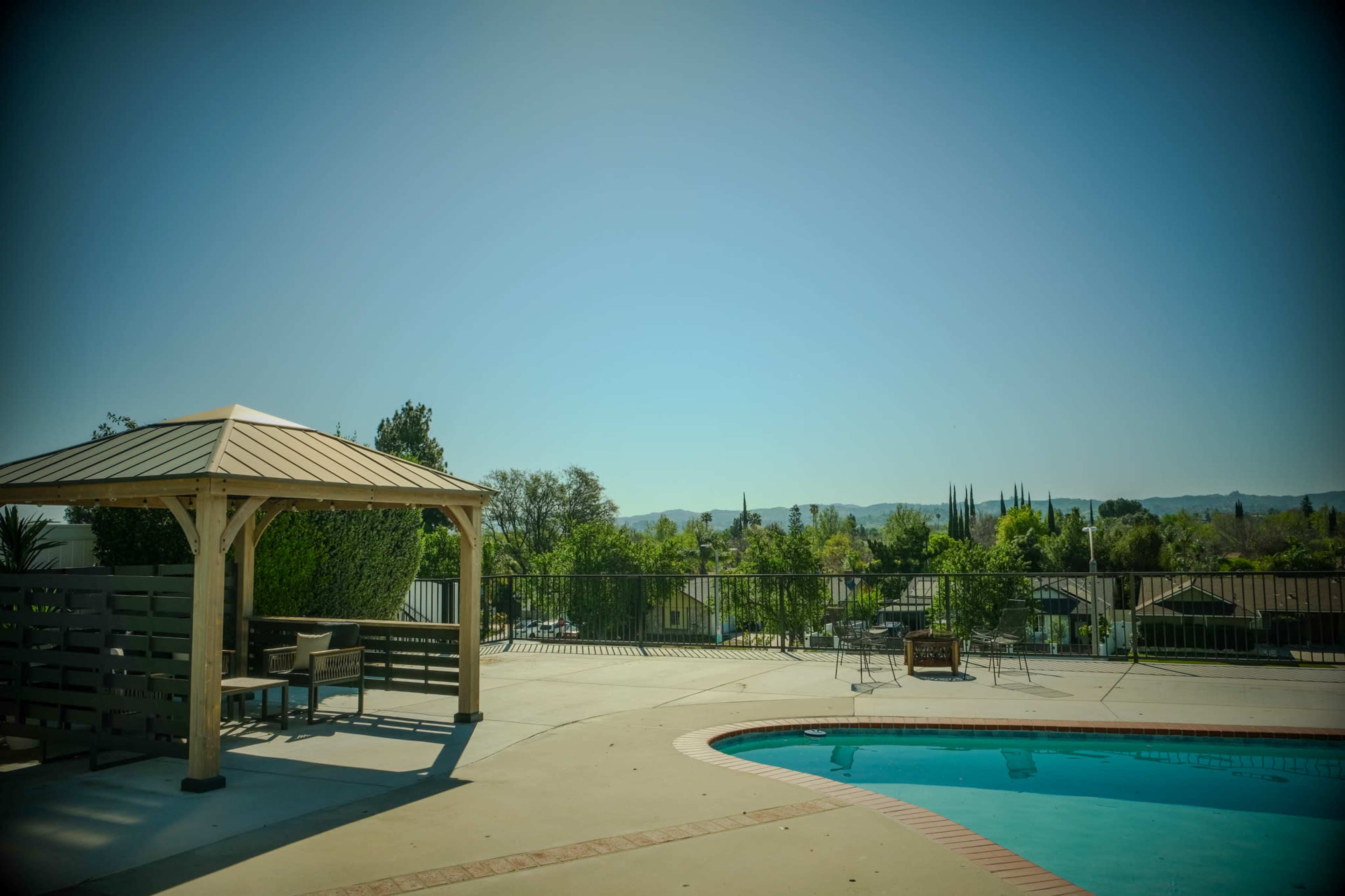 A gazebo stands beside a swimming pool, overlooking a landscape of trees and distant hills under a clear blue sky.