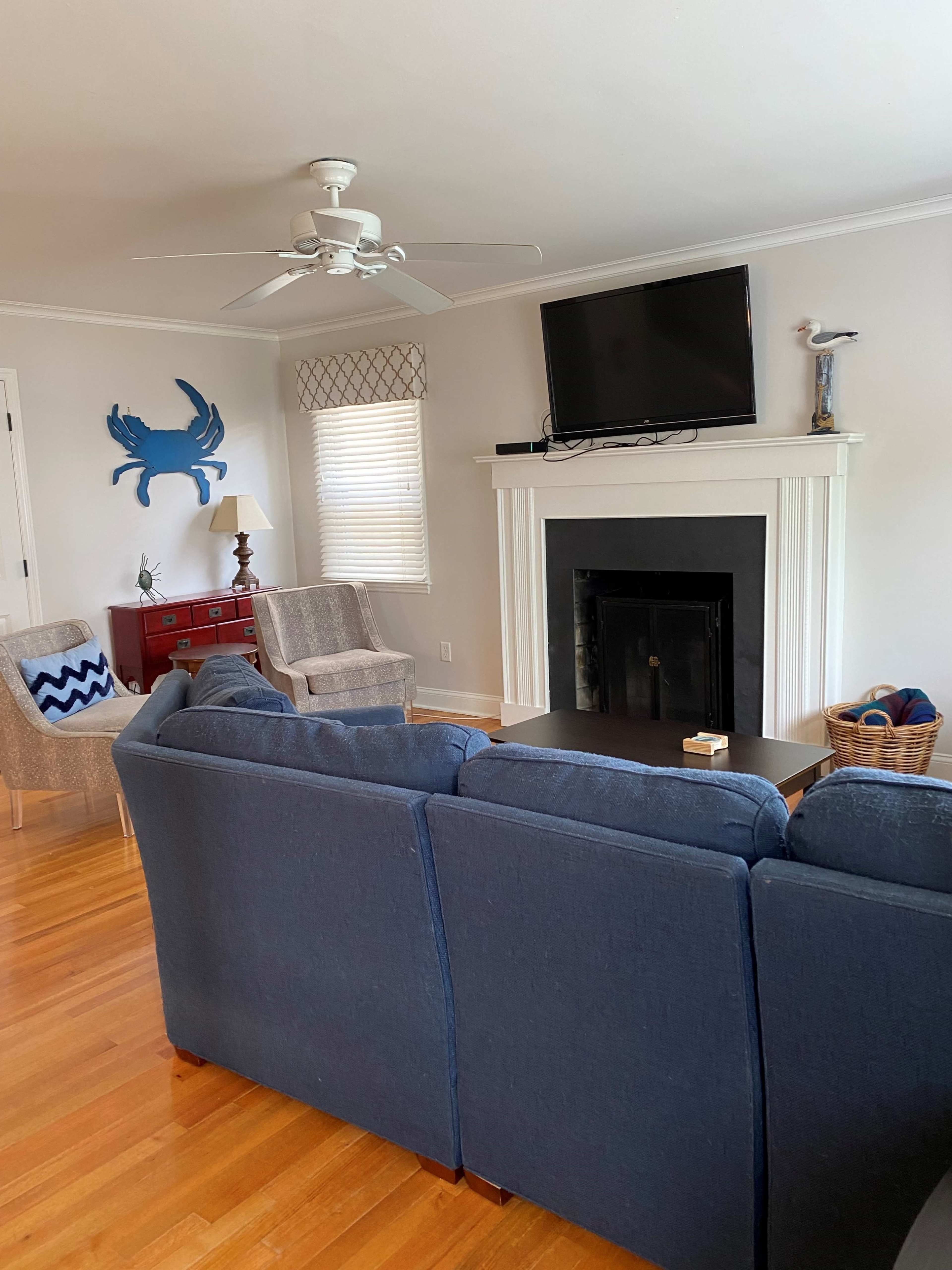 A living room with a dark blue couch facing a coffee table, a fireplace with a television mounted above it, and two armchairs in front of the fireplace.