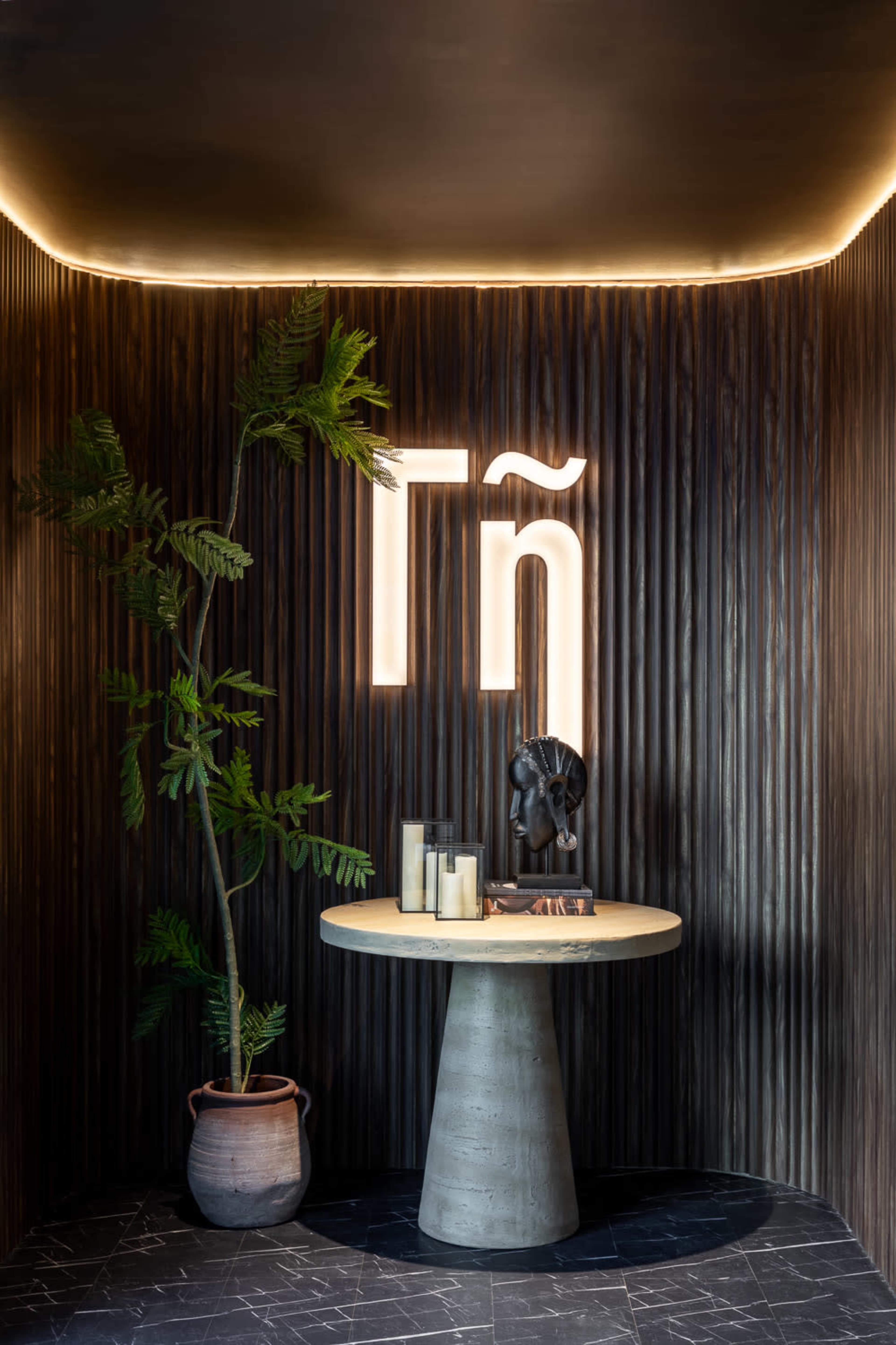 The image shows a modern reception area featuring a round concrete table, a decorative plant in a clay pot, and backlit text on wood paneling.