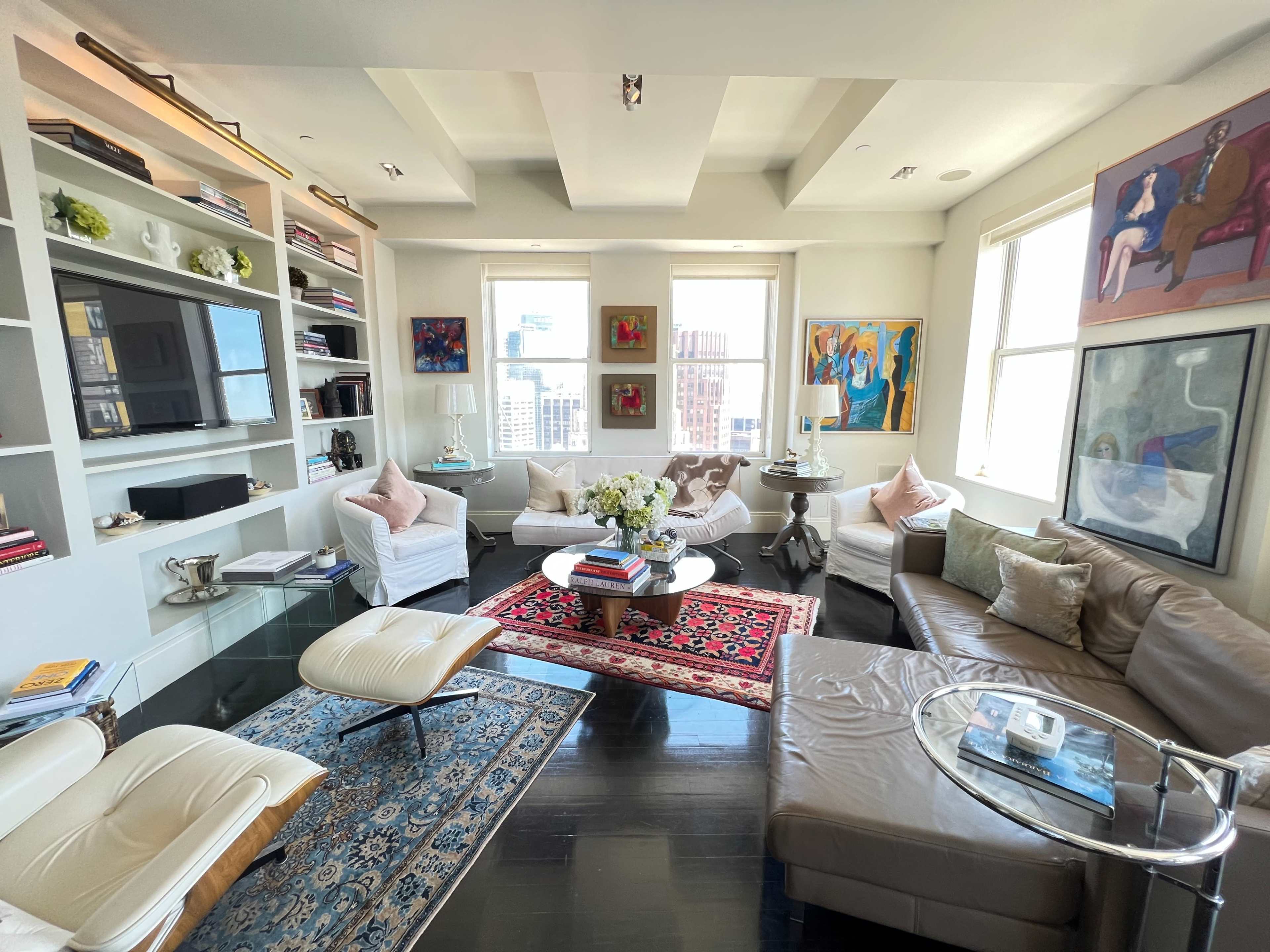 A modern living room with comfortable seating, an area rug, bookshelves, and large windows providing a view of the city.