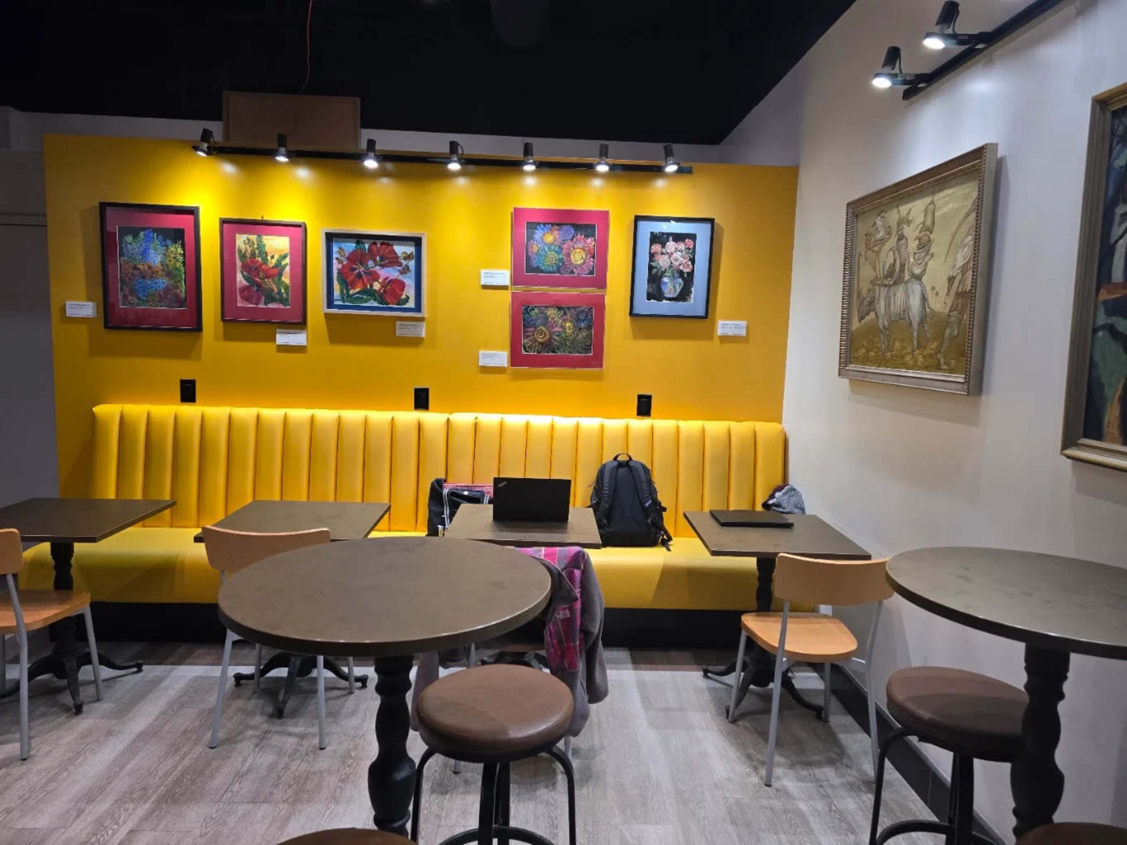 The image shows a cozy cafe interior with a bright yellow wall adorned with framed artwork, featuring a long cushioned seating area and several round tables.