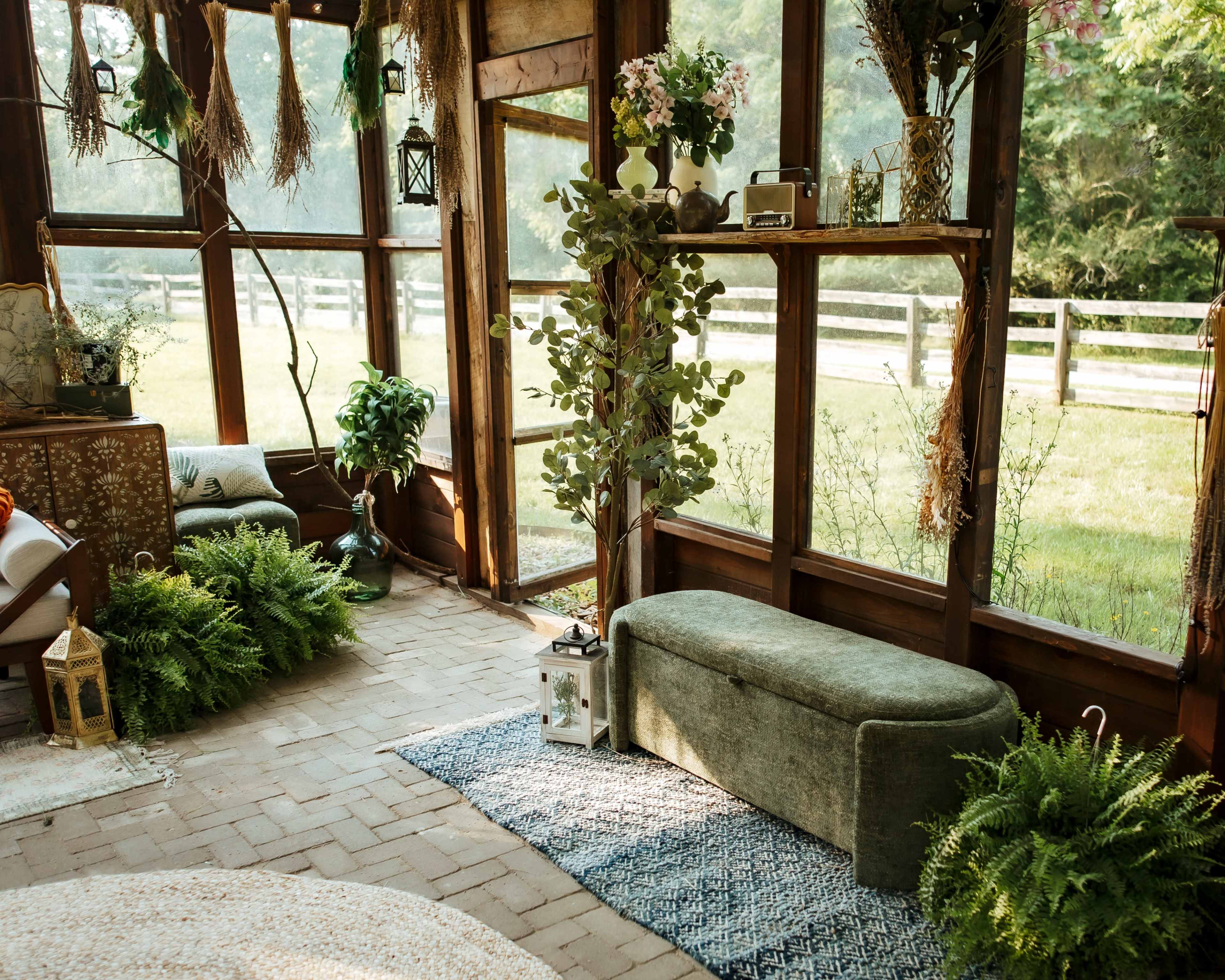 The image shows a sunlit sunroom filled with greenery, a bench, and decorative items arranged on wooden shelves.