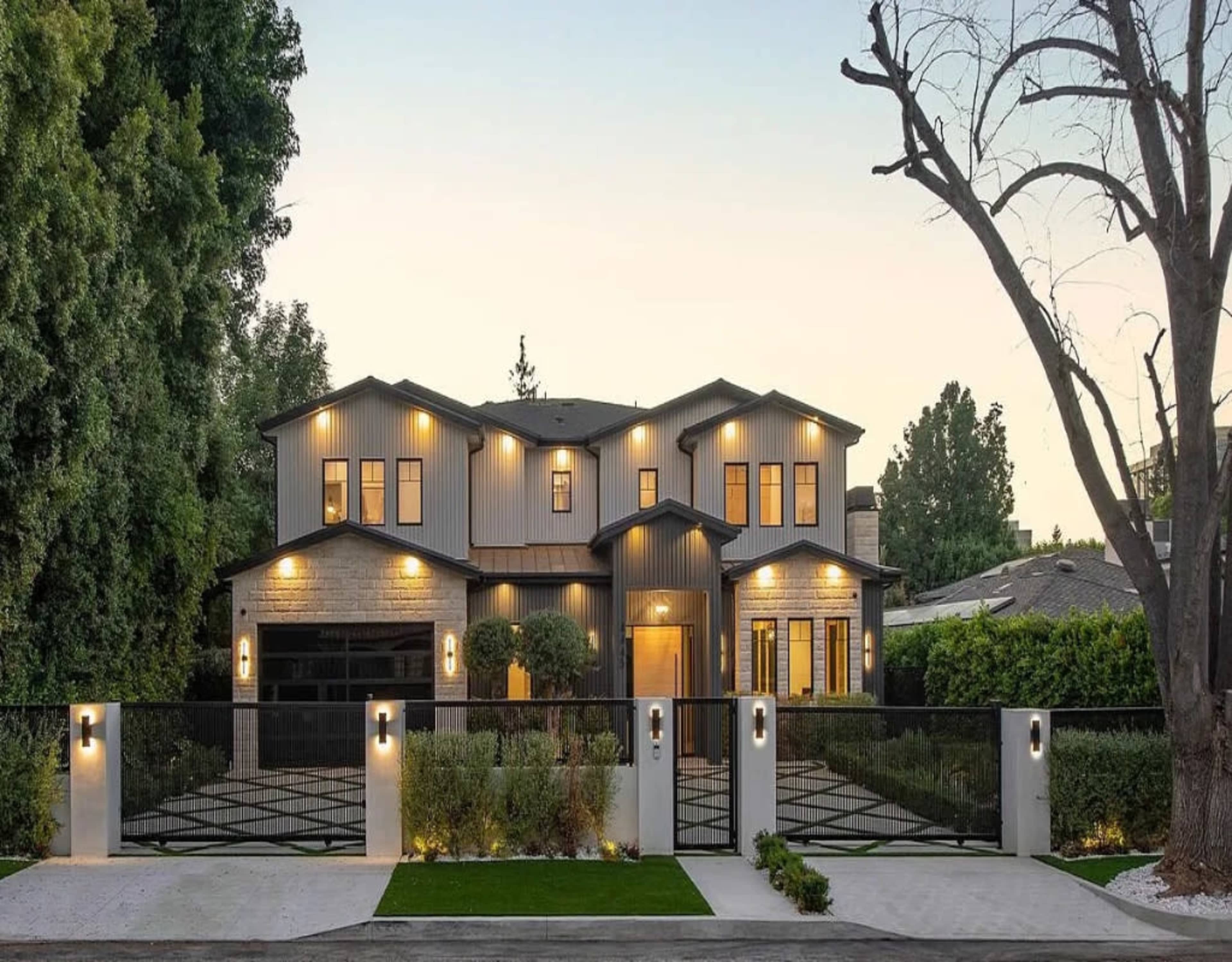 The image shows a modern two-story house with a mix of stone and metal siding, surrounded by greenery and featuring a gated entrance with landscaped pathways.