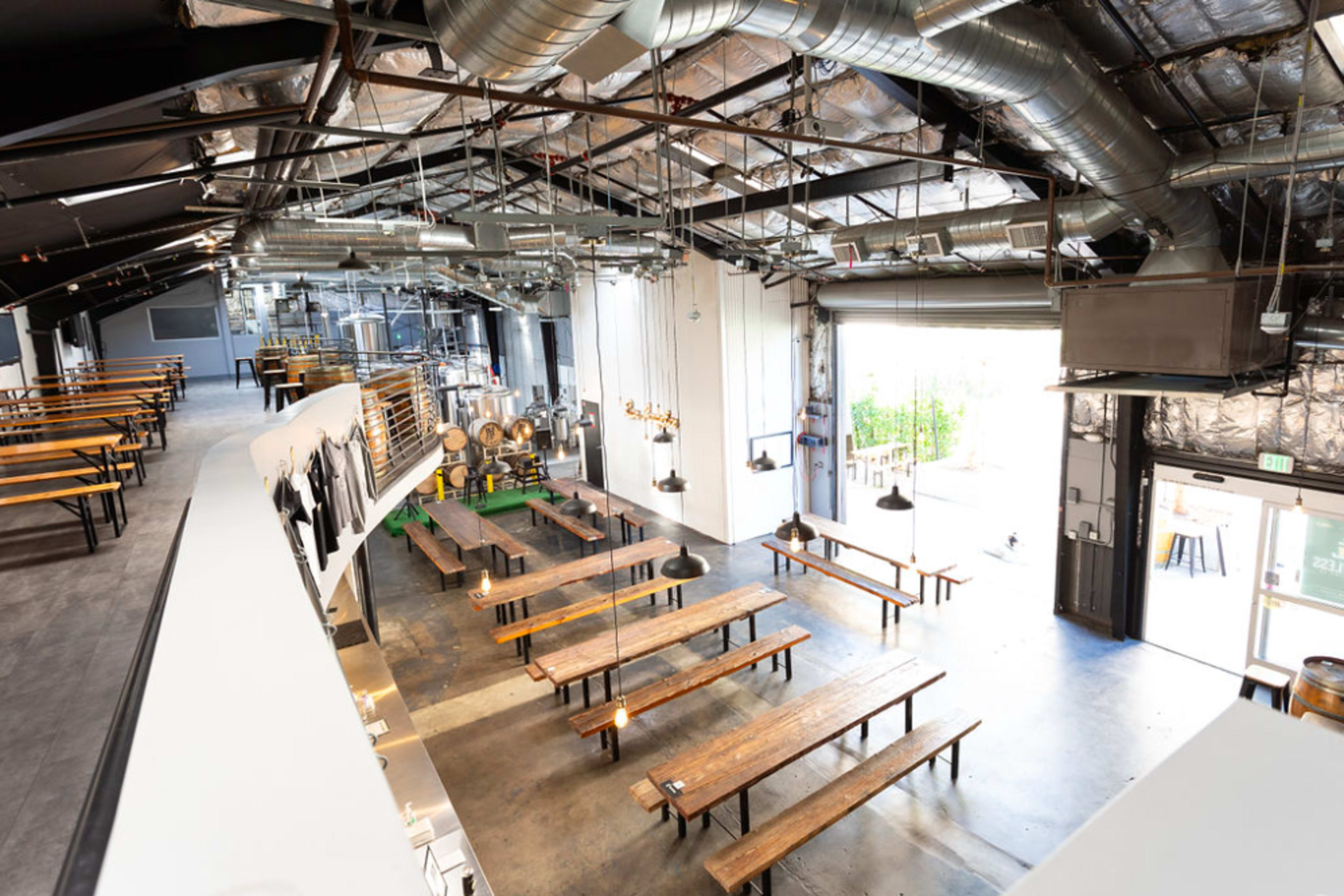 A spacious industrial-style restaurant interior with wooden communal tables, high ceilings, and large windows letting in natural light.