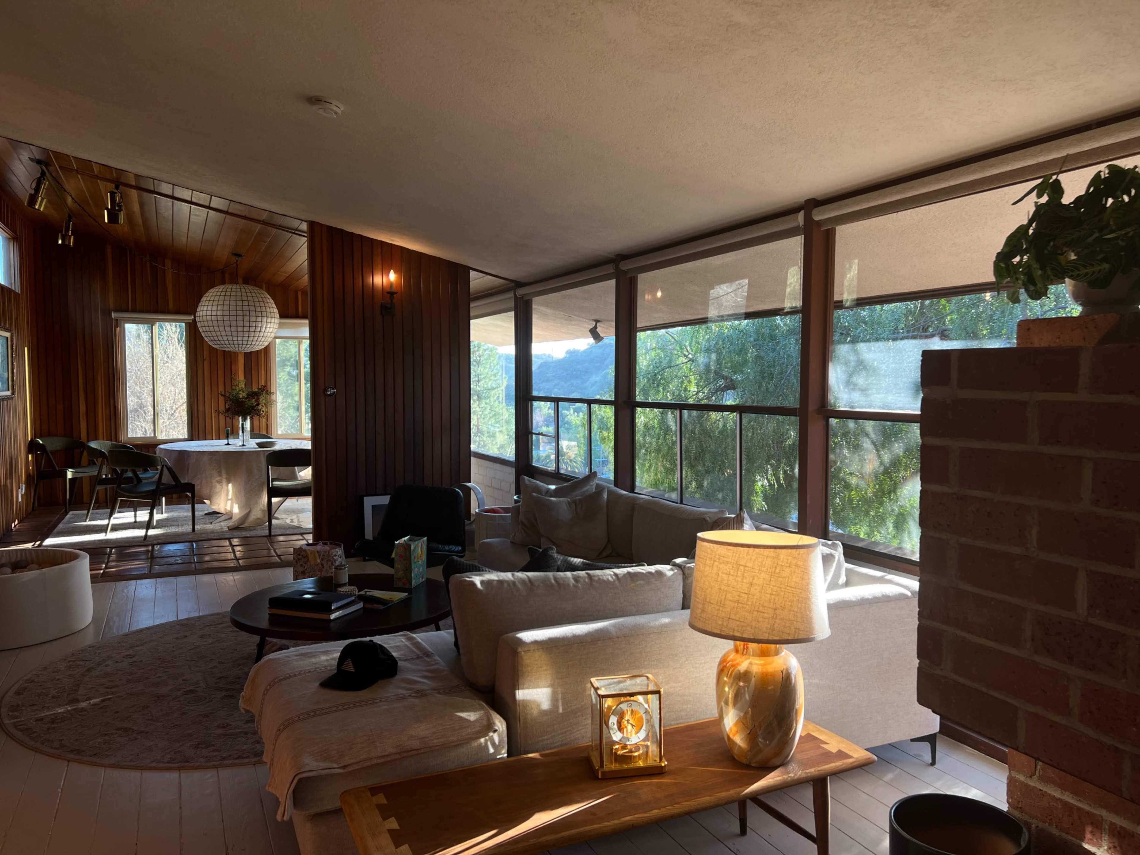 The image shows a modern living room with large windows overlooking a natural landscape, featuring a sofa, a coffee table, and a dining area beyond.