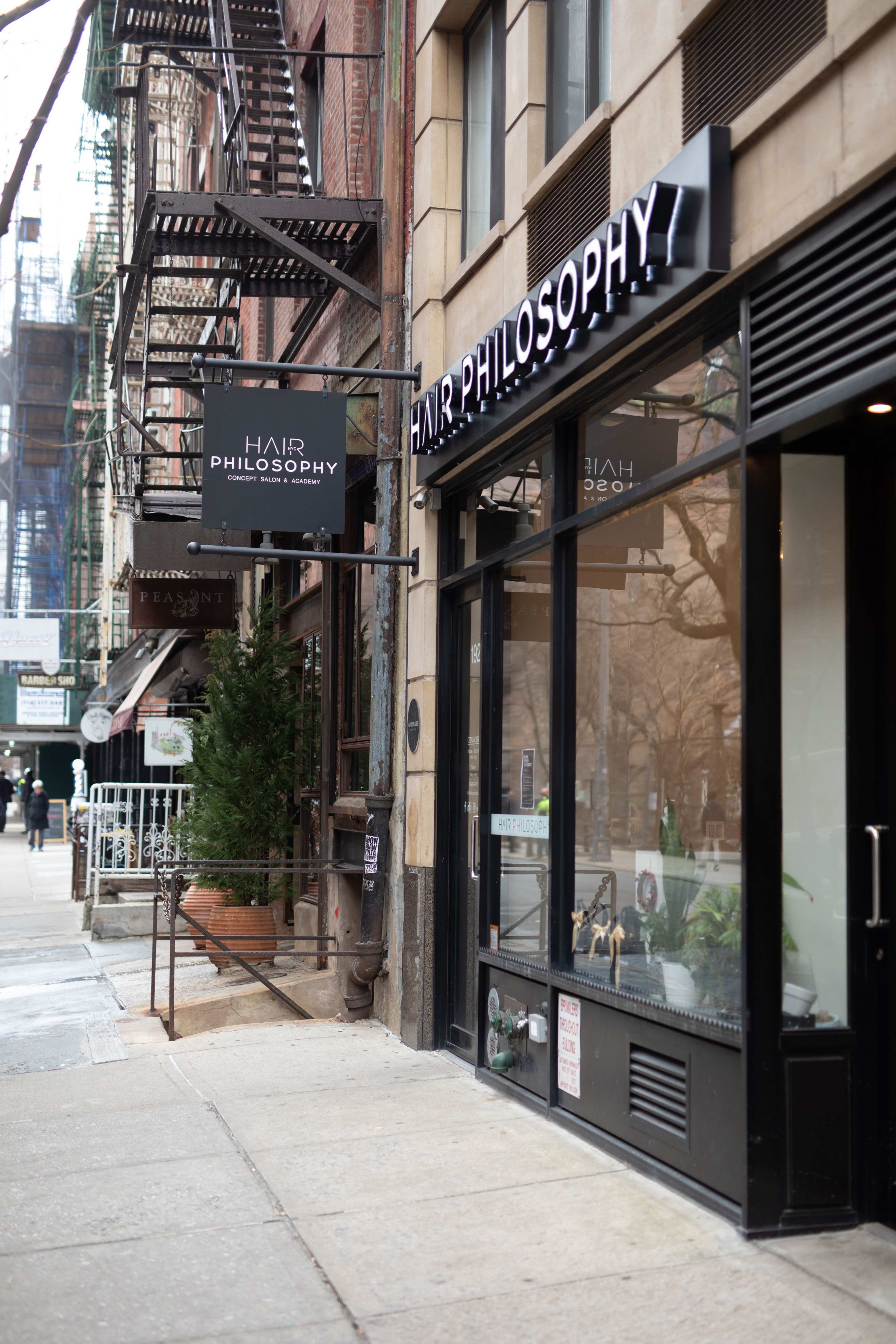 A street view of a hair salon named "Hair Philosophy," with a black sign hanging above its entrance and decorative plants outside.