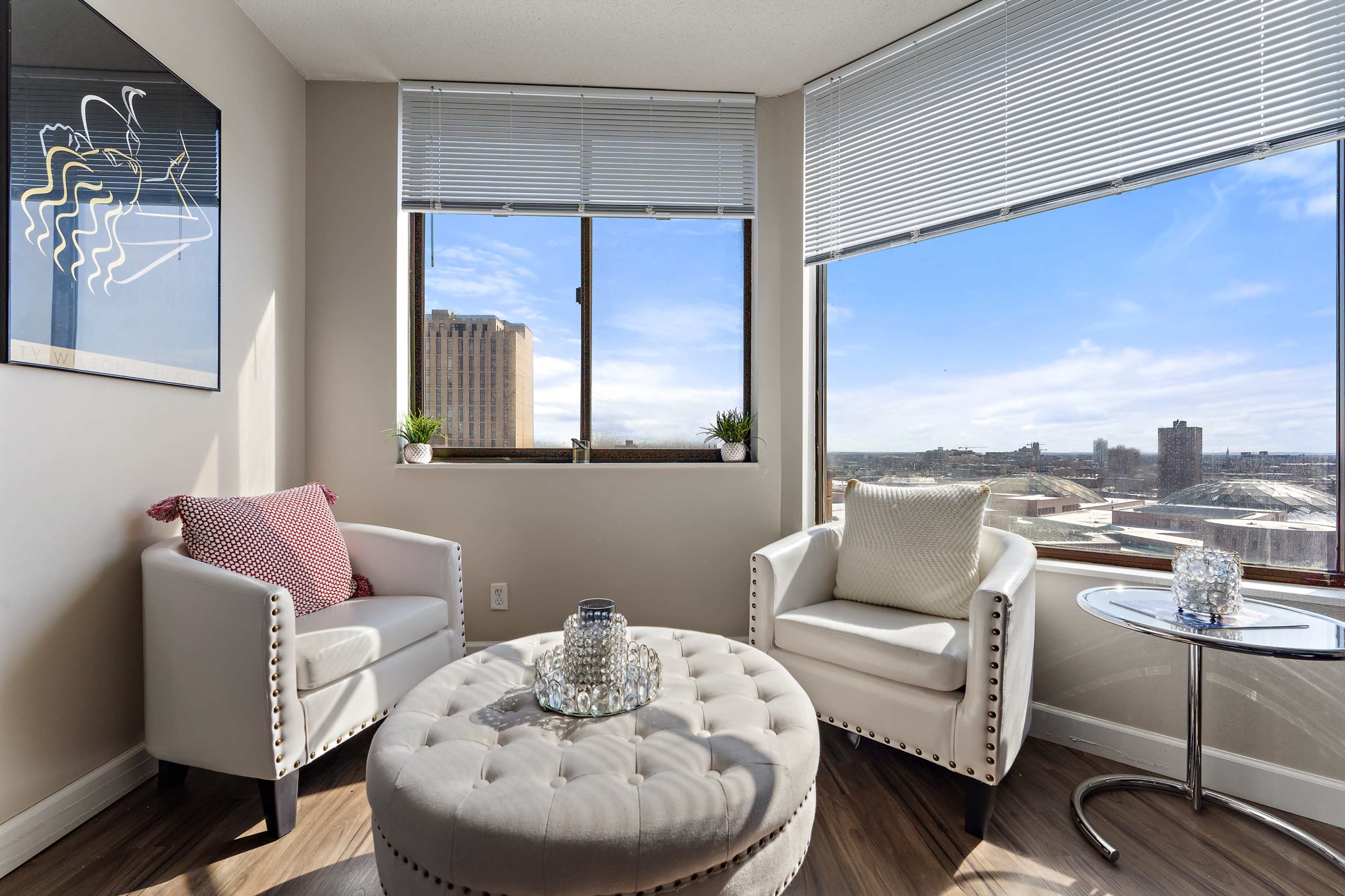 A bright corner of a room featuring two white armchairs, a round ottoman, and a small side table, with large windows offering a view of the city skyline.