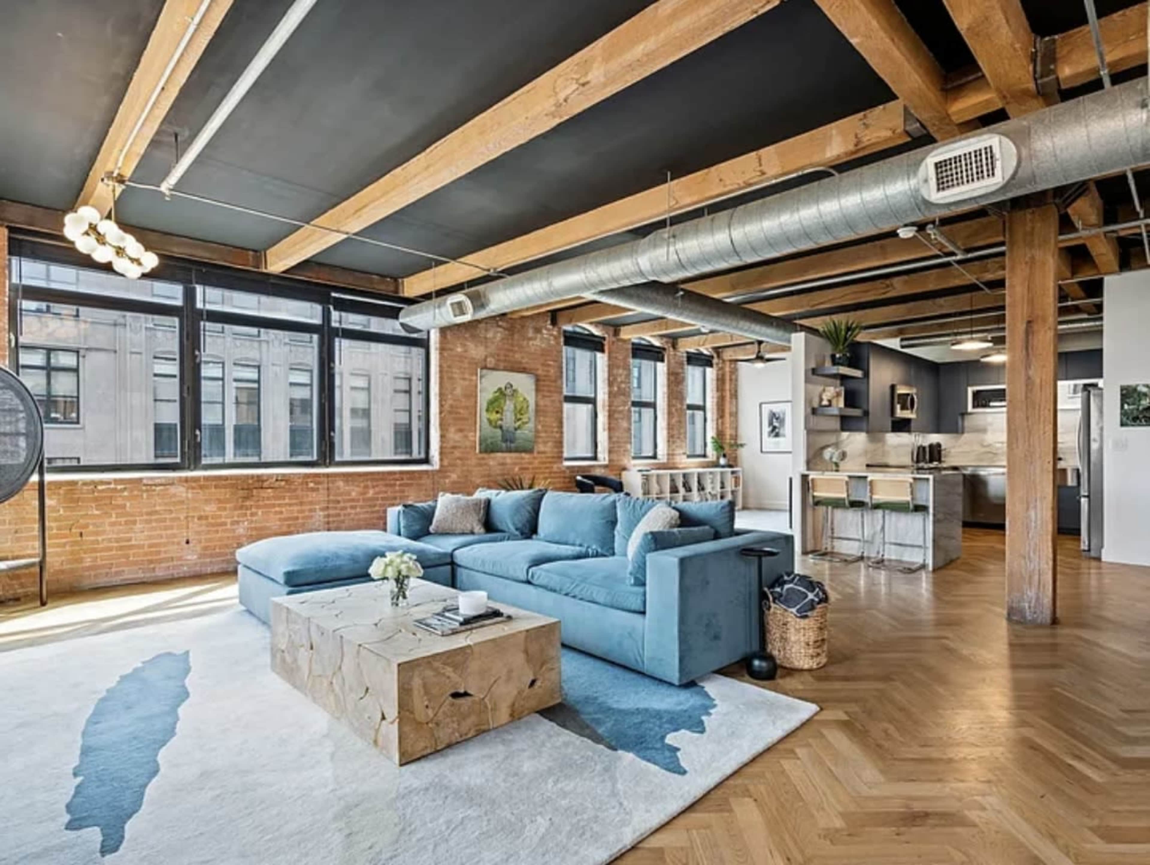 The image shows a modern living room with a blue sectional sofa, hardwood floors, exposed brick walls, and large windows allowing natural light.
