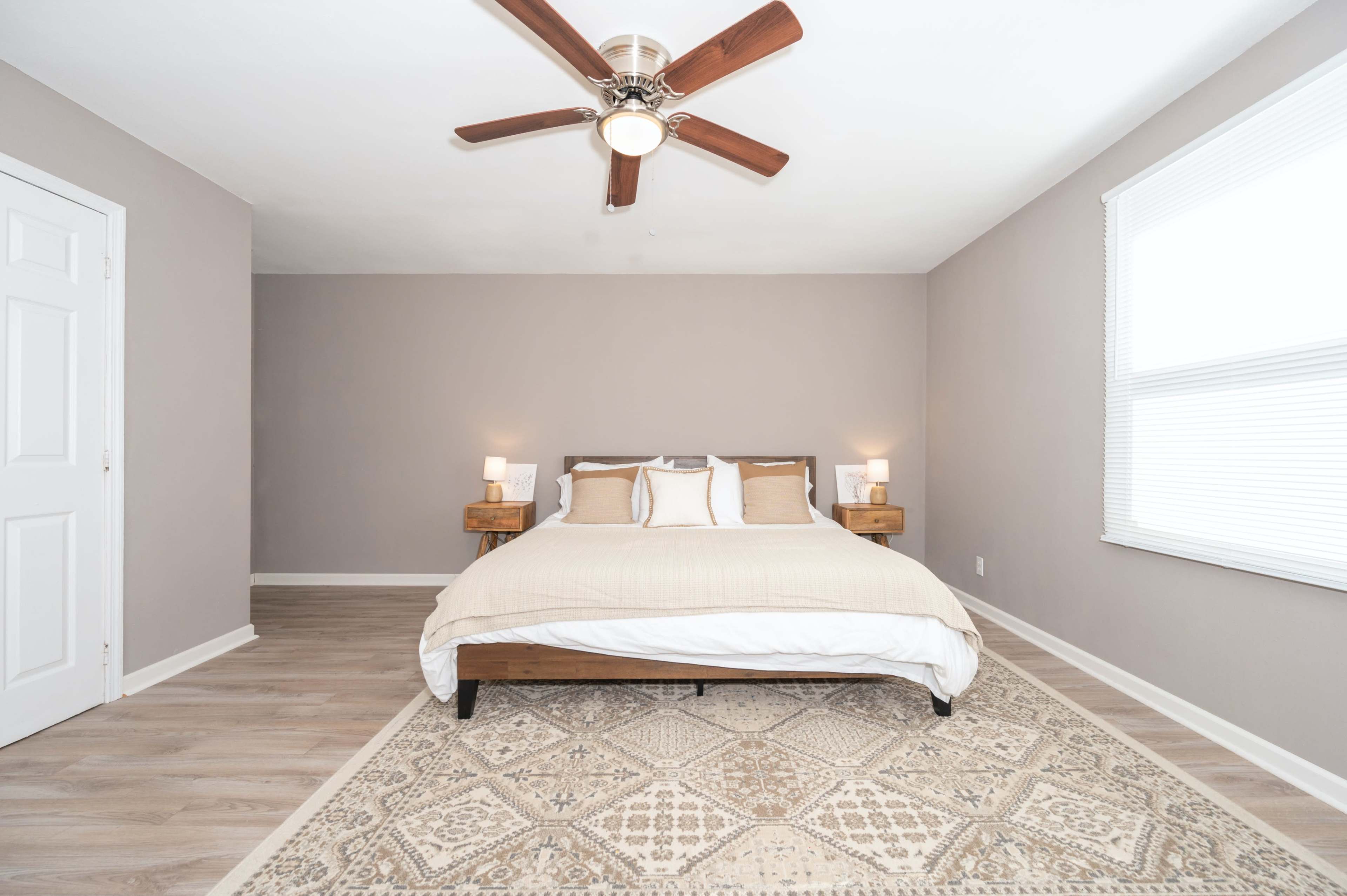 A minimalist bedroom featuring a large bed with beige bedding, two bedside lamps, and a patterned rug on a wooden floor.