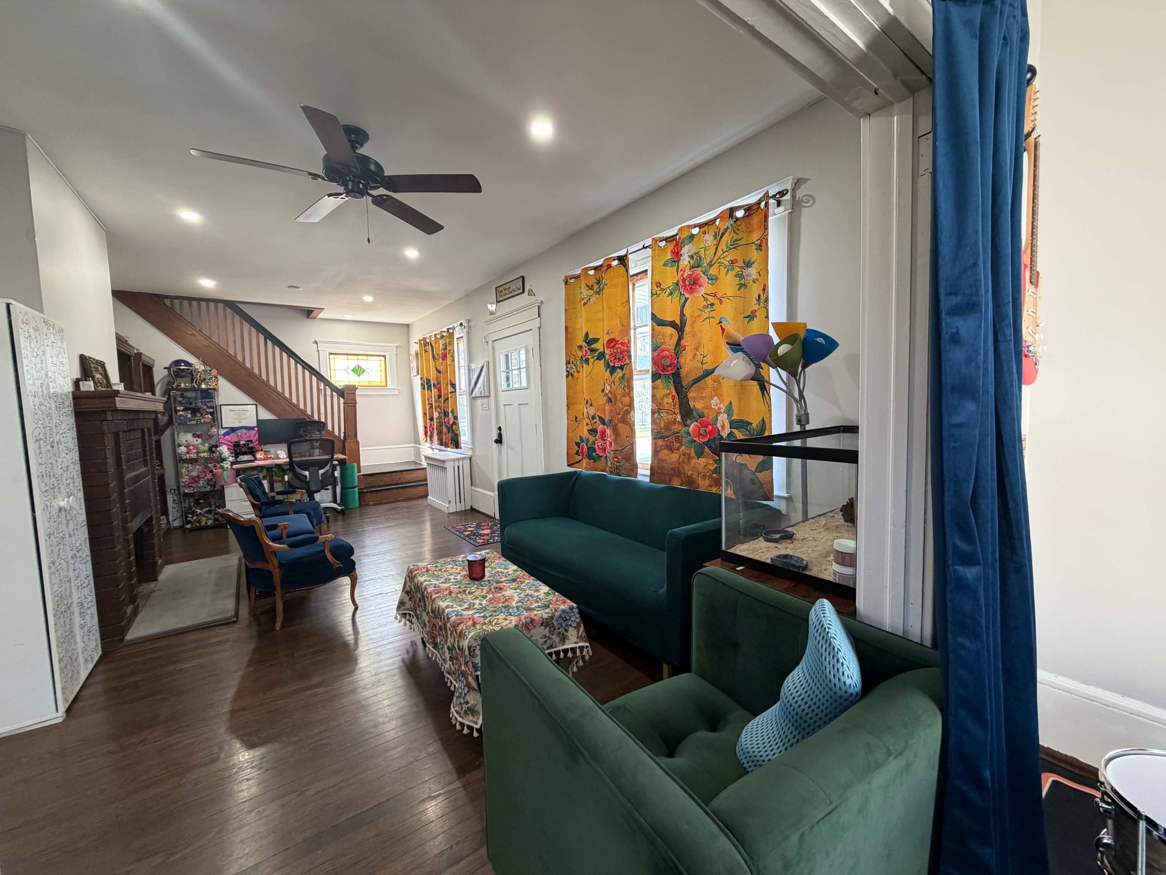 The image shows a living room with green sofas, a colorful tablecloth on a coffee table, bright floral curtains, and wooden flooring.