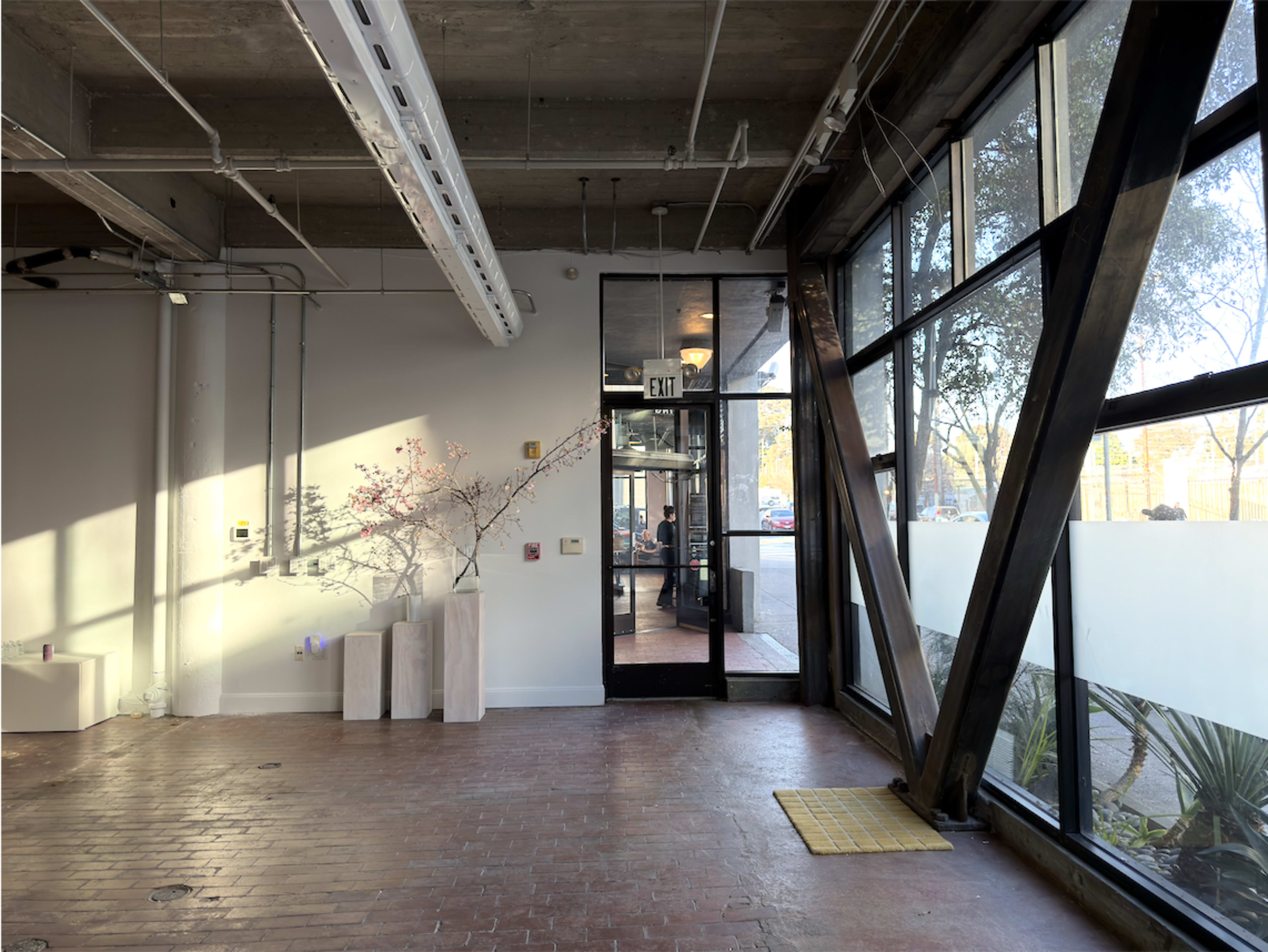 A spacious interior features large windows, exposed beams, and minimal furnishings, leading to an exit door that opens to an outdoor area.