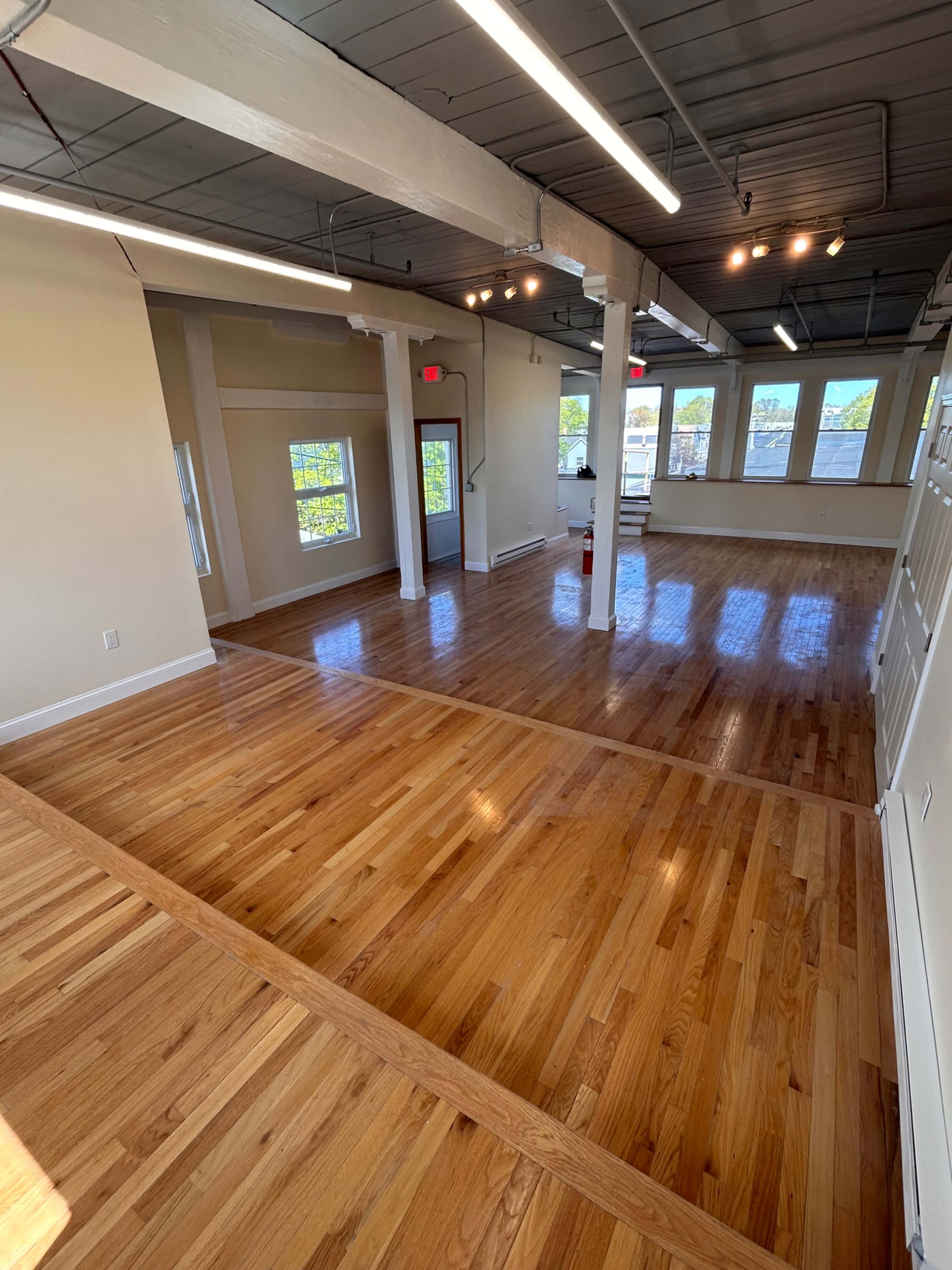 The image shows a spacious, modern interior with polished wooden floors, large windows, and exposed beams.