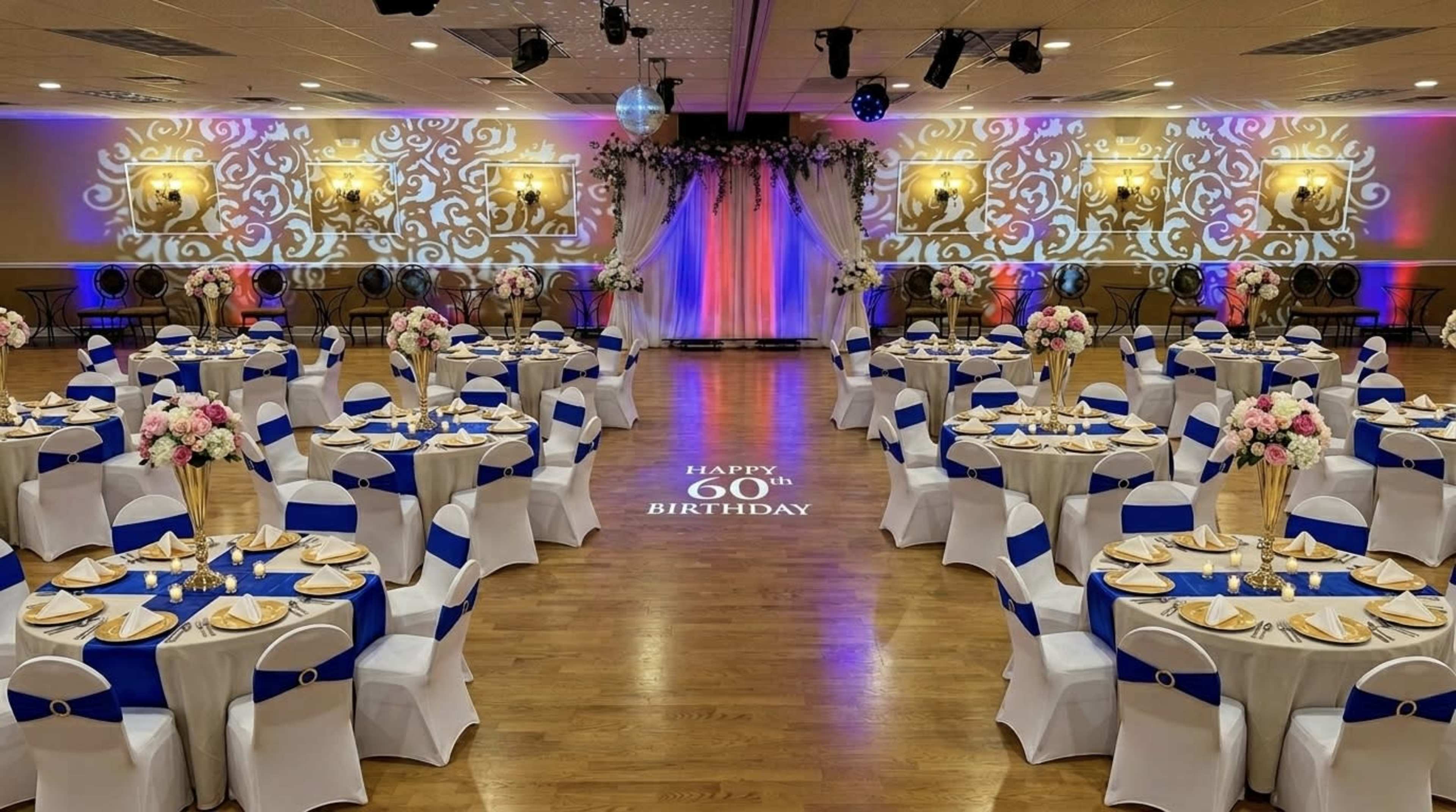 The image shows a decorated banquet hall set up for a 60th birthday celebration, featuring multiple tables with floral centerpieces and blue accents, as well as a backdrop with the birthday message.